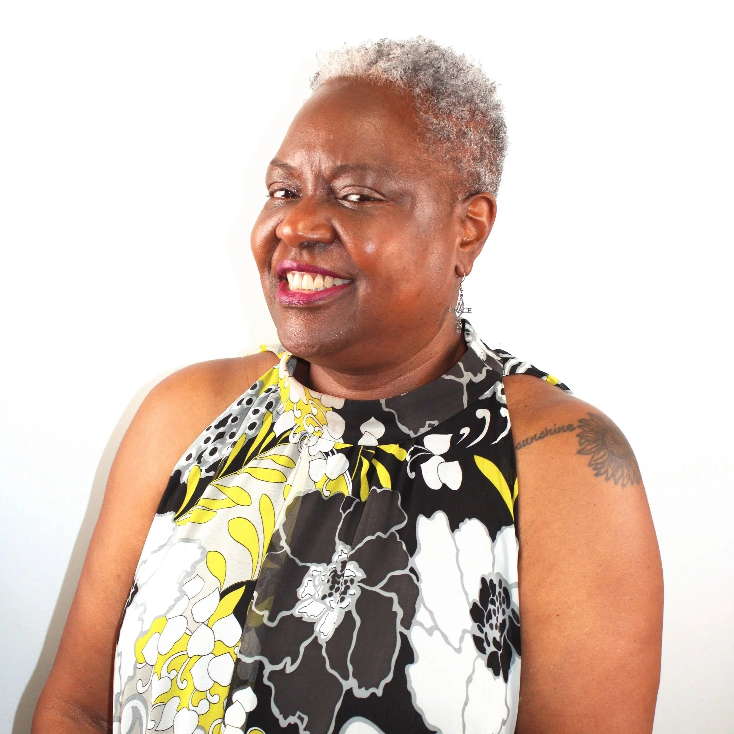 Portrait of a smiling woman with short gray hair, wearing a colorful floral sleeveless top, standing against a white background.