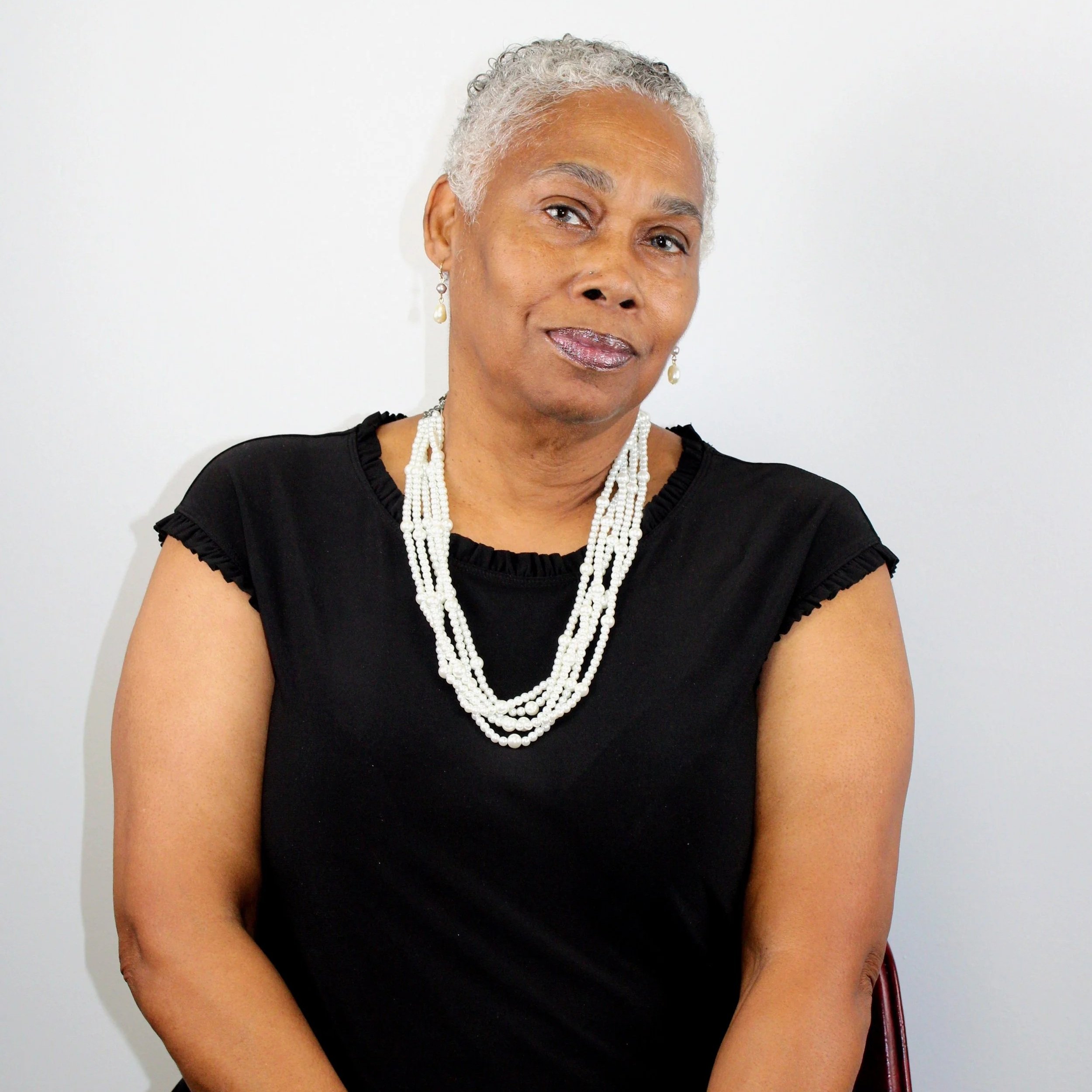 An older woman with short gray curly hair wearing a black short-sleeve top and multiple strands of white pearls, smiling softly against a plain white background.