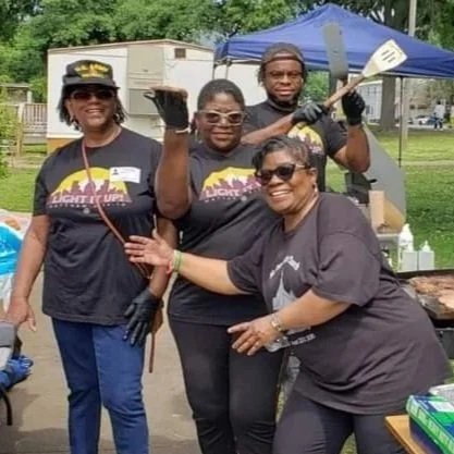 Group of four women and one man smiling outdoors at a barbecue event, some wearing matching T-shirts and one woman holding a spatula.