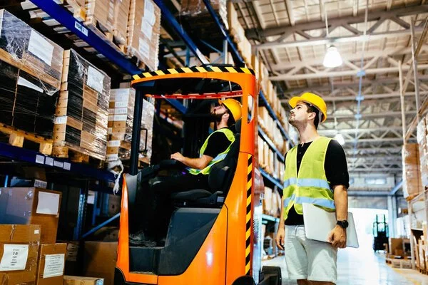 Two workers in a warehouse, one operating a forklift and the other inspecting or supervising. Both wear safety helmets and vests.