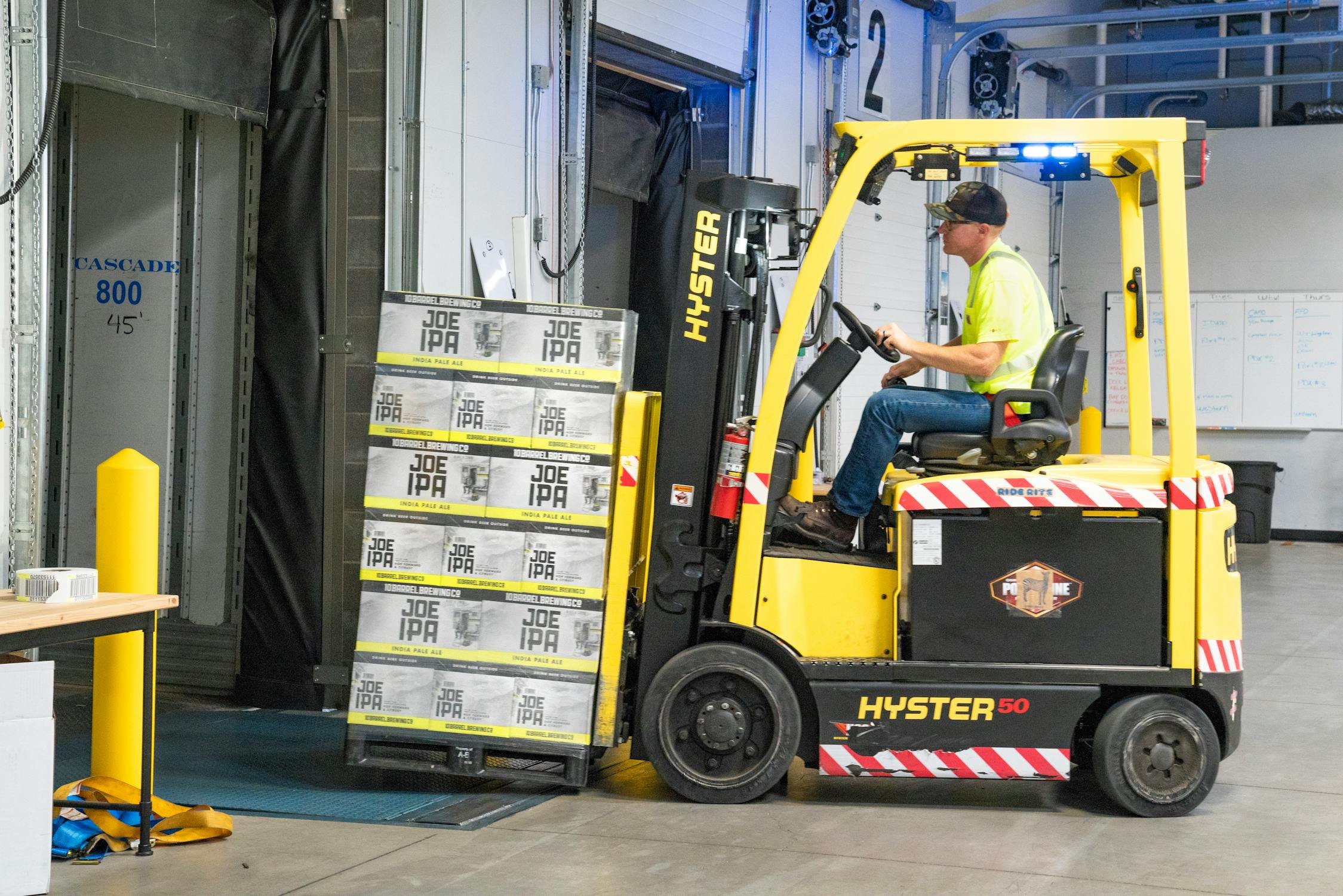 A man operating a yellow Hyster forklift truck in a warehouse, lifting a pallet of boxed Joe IPA India Pale Ale beer bottles.