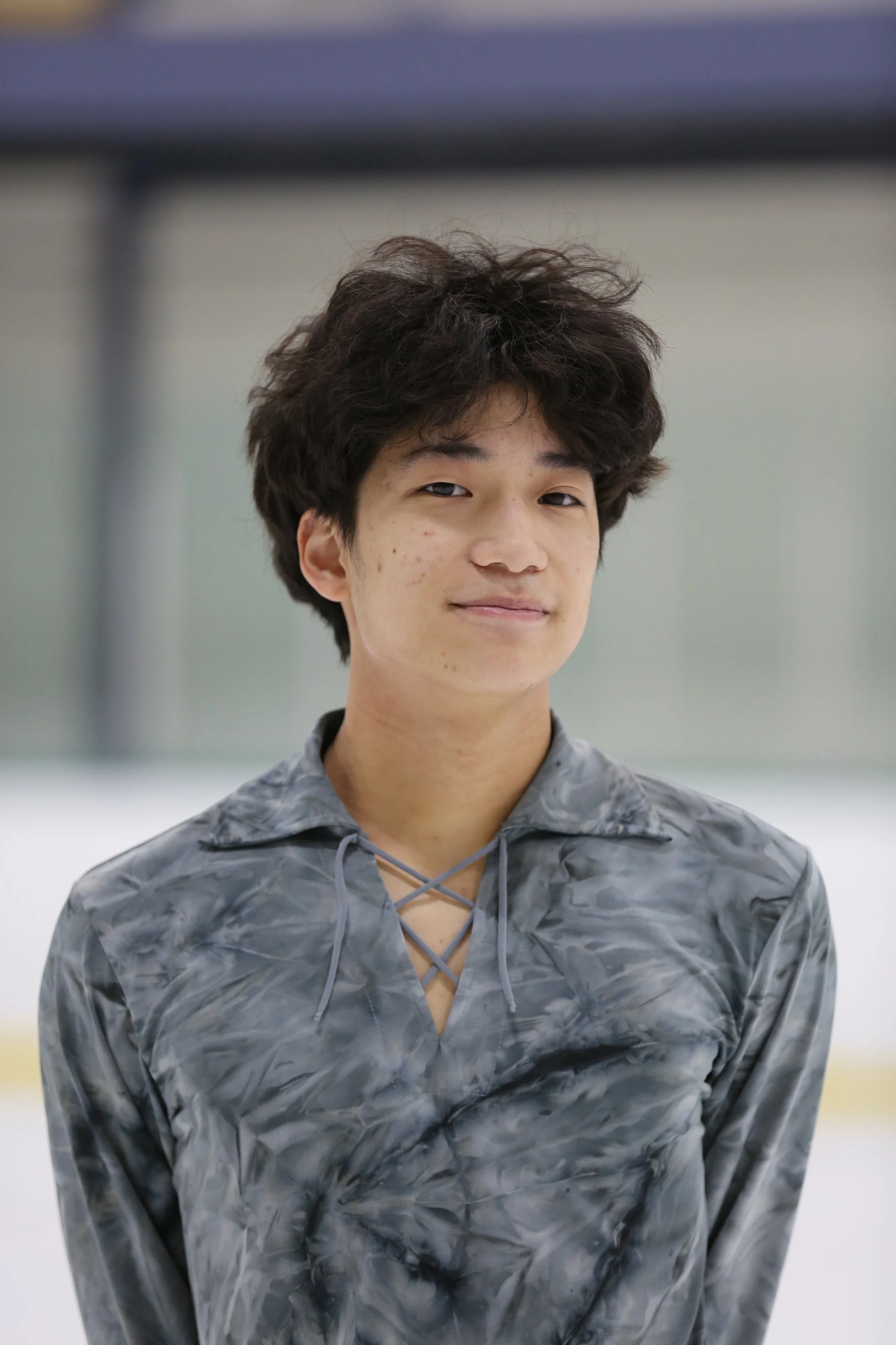 A young man with dark curly hair smiling at the camera, wearing a white figure skating costume with rhinestones, standing on an ice rink.