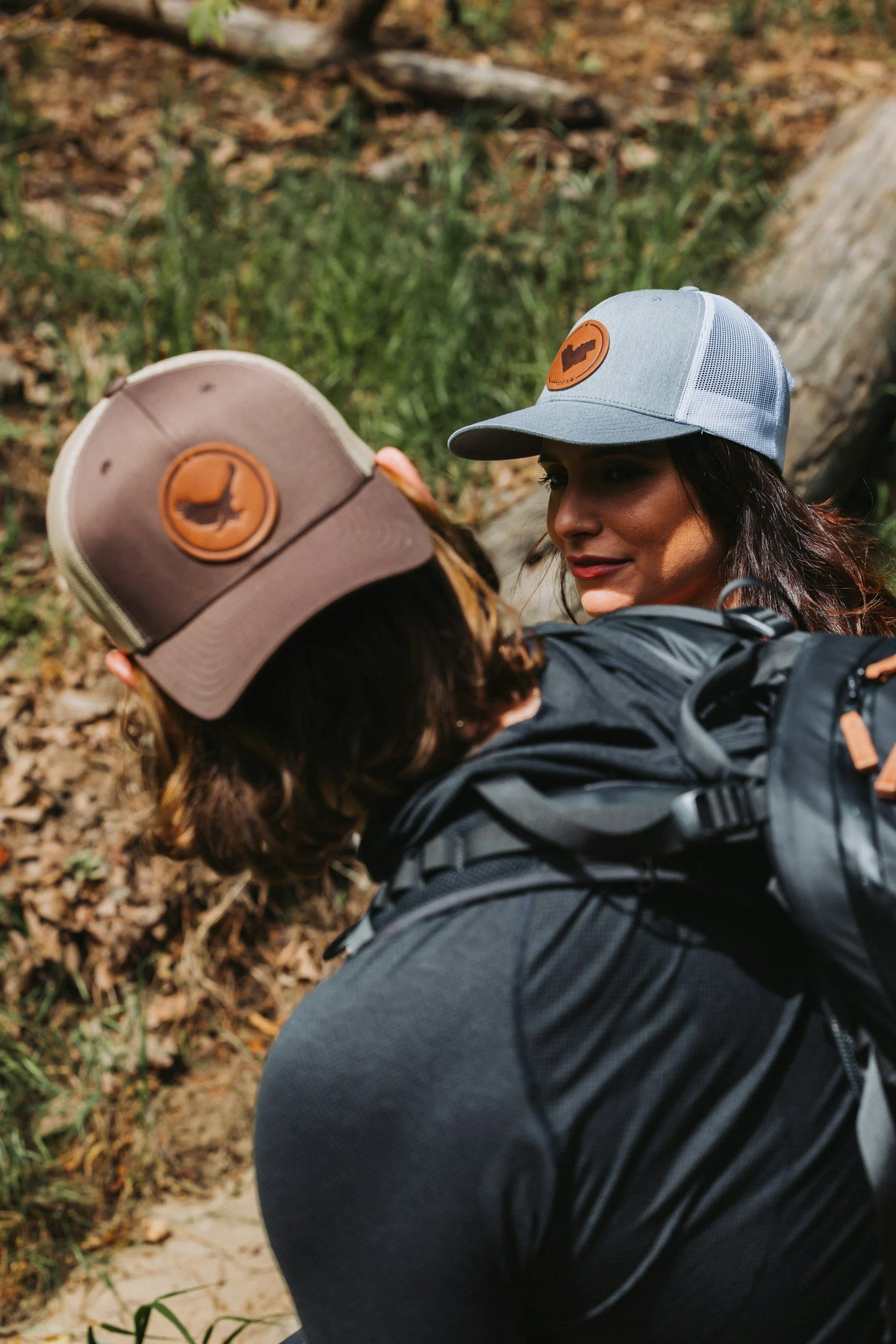 Two women wearing caps with patch designs, one in grey and the other in beige, outdoors in a wooded area.
