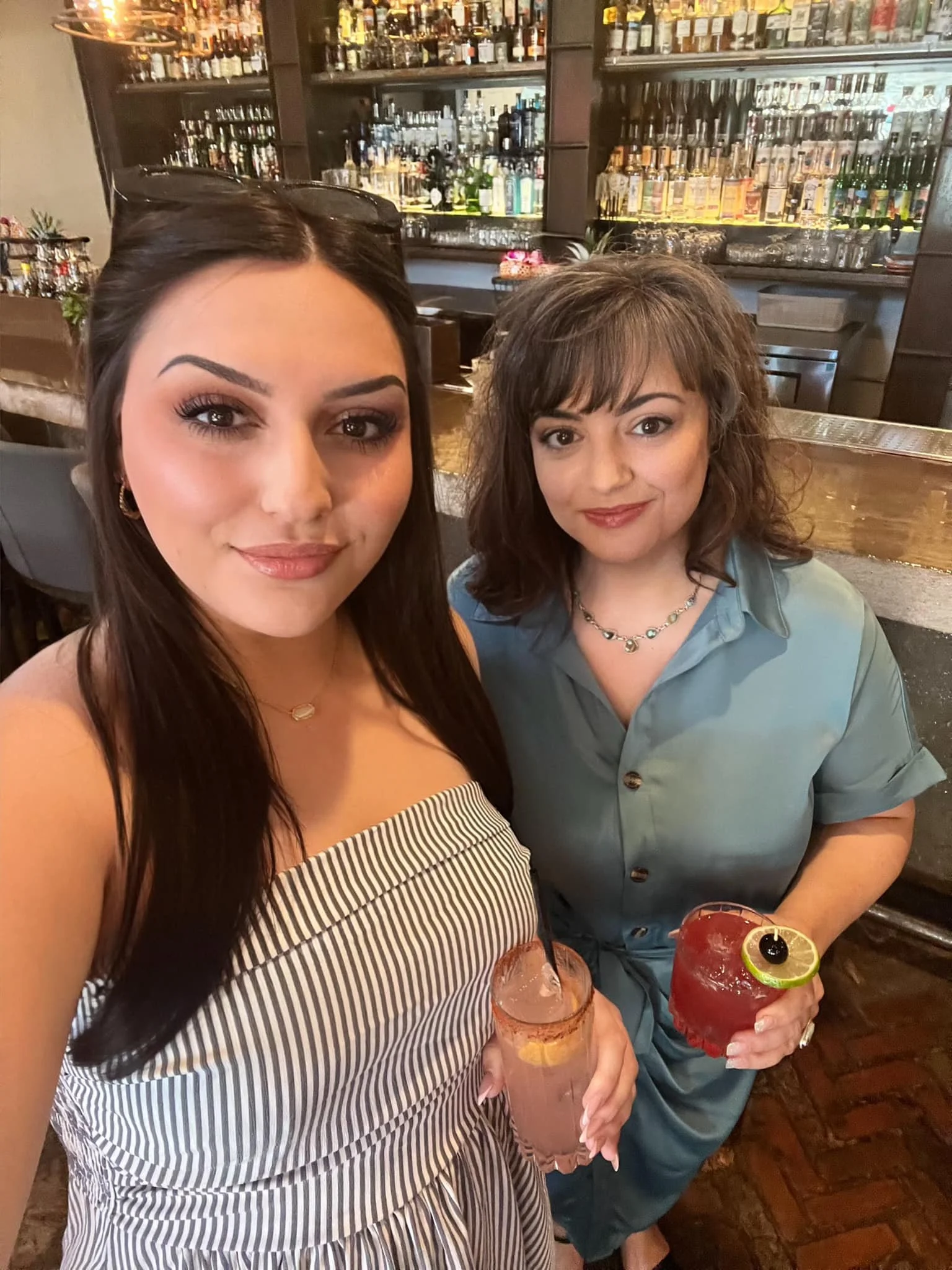 Two women taking a selfie at a bar, each holding a colorful cocktail. The bar in the background has multiple bottles of alcohol displayed on shelves.