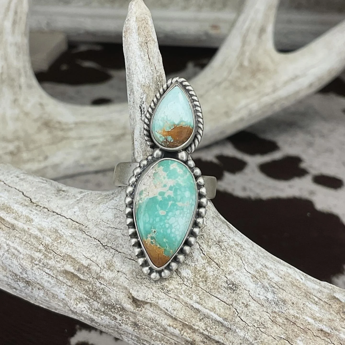 Silver ring with two turquoise stones, one in a teardrop shape and the other in an elongated oval shape, resting on a weathered antler with a wooden background.