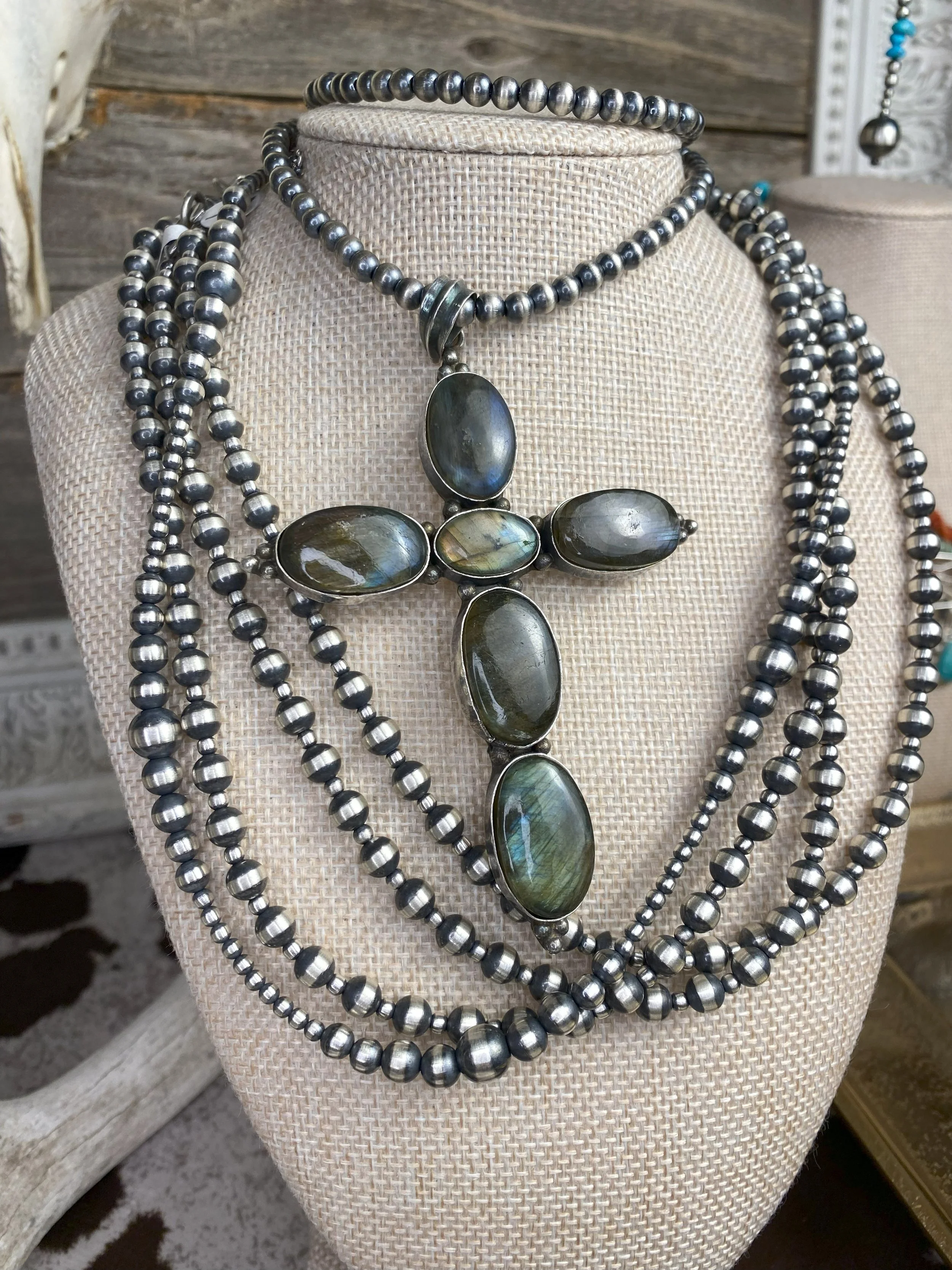 A display of multiple layered silver beaded necklaces, with one featuring a large cross pendant made of iridescent oval stones.