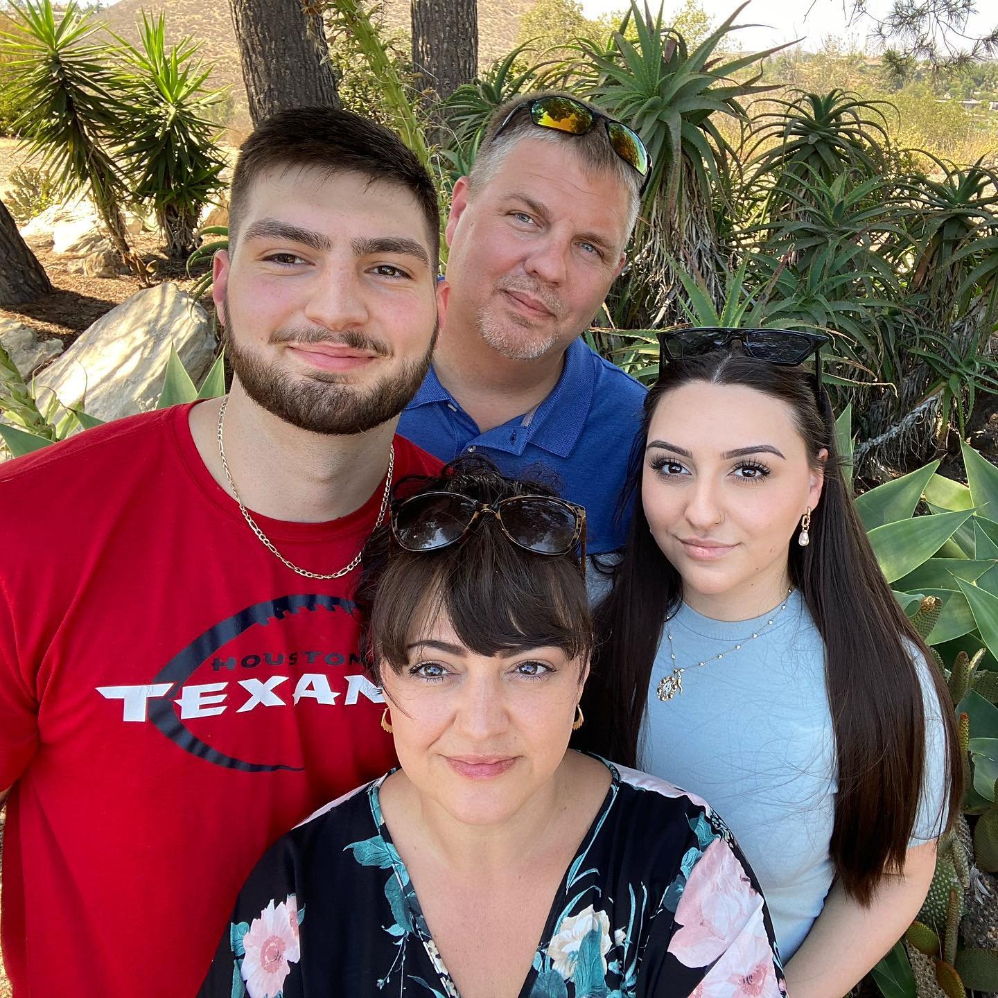 Group of five people posing outdoors among green plants and trees, smiling at the camera.
