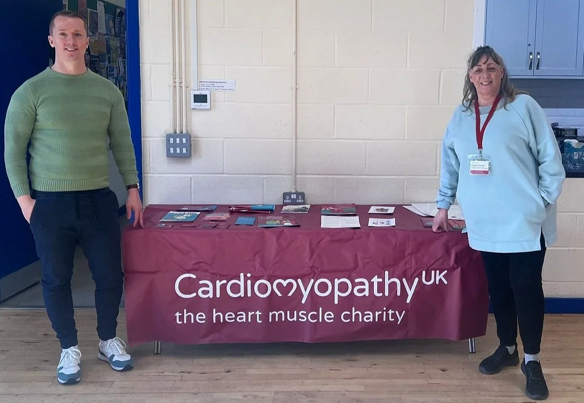 Two people standing beside a table with a maroon tablecloth displaying information about Cardiology UK, a heart muscle charity, in a community center or similar indoor setting.