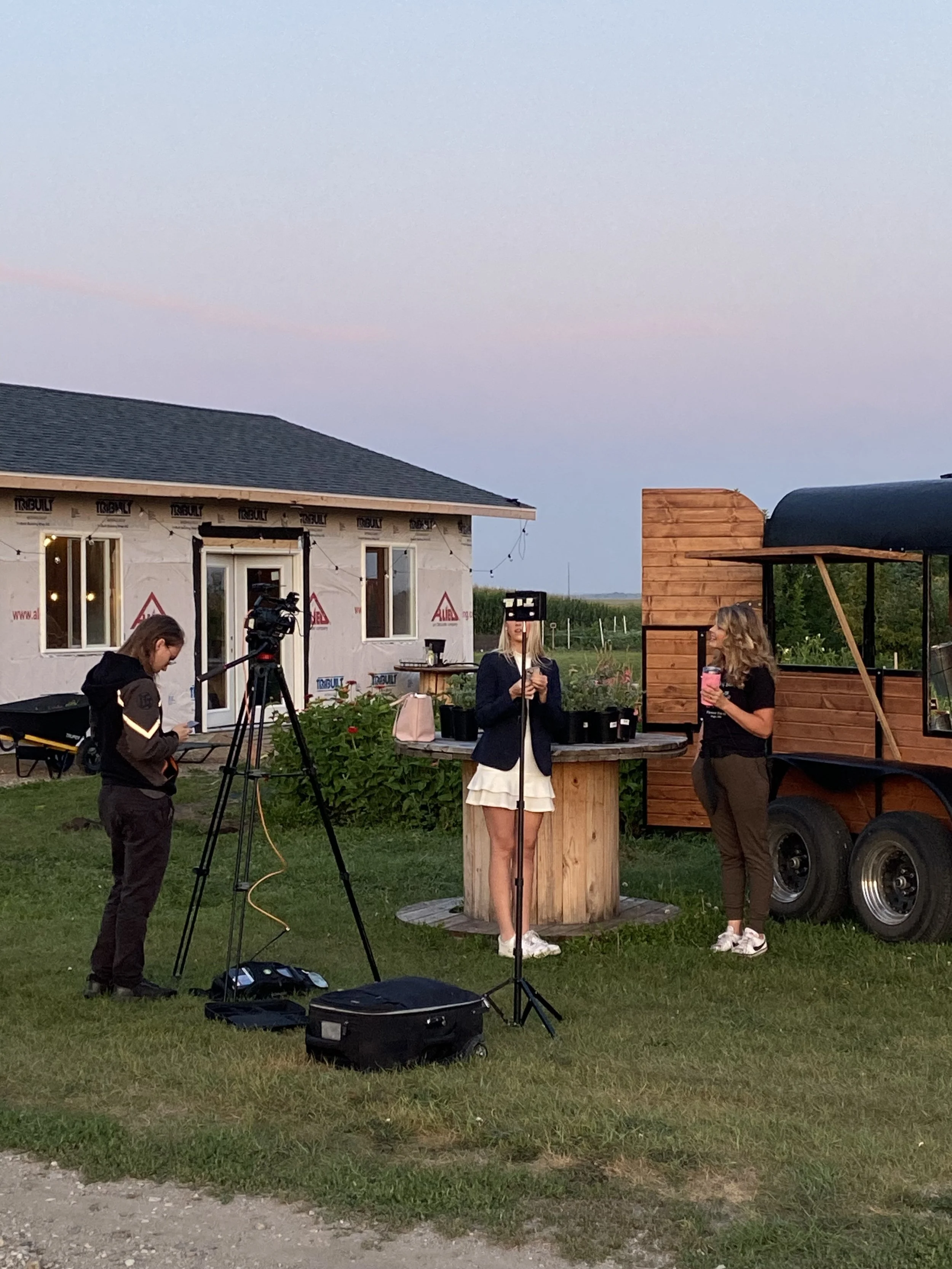 A woman speaking into a microphone at an outdoor event with a camera operator recording, a woman holding a drink, and a food trailer in the background during sunset.
