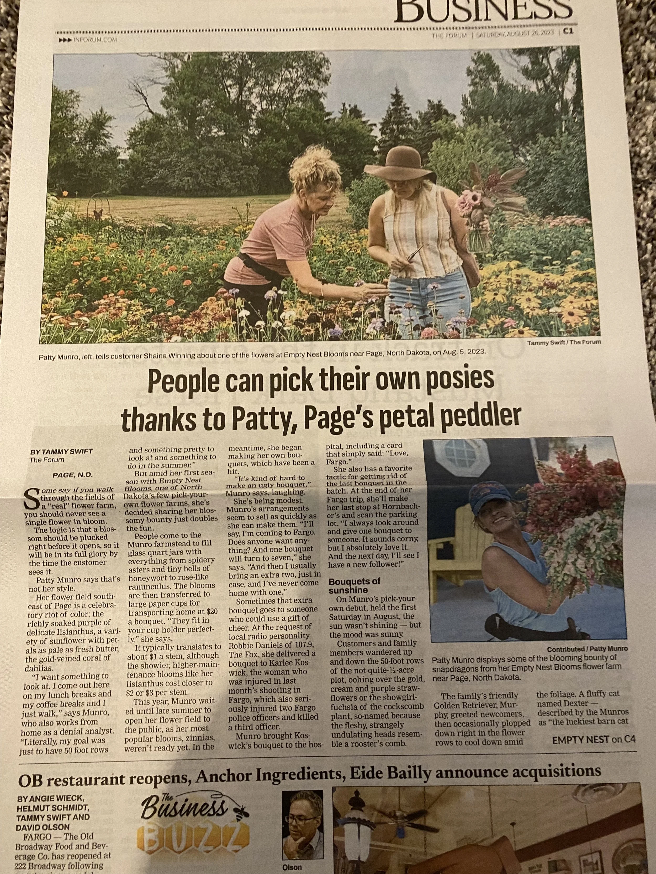 A newspaper story with a photo of two women in a flower field. One woman, wearing a pink shirt, shows a flower to the other woman who is wearing a striped sleeveless top and a large hat. The field is filled with colorful flowers with trees in the bac