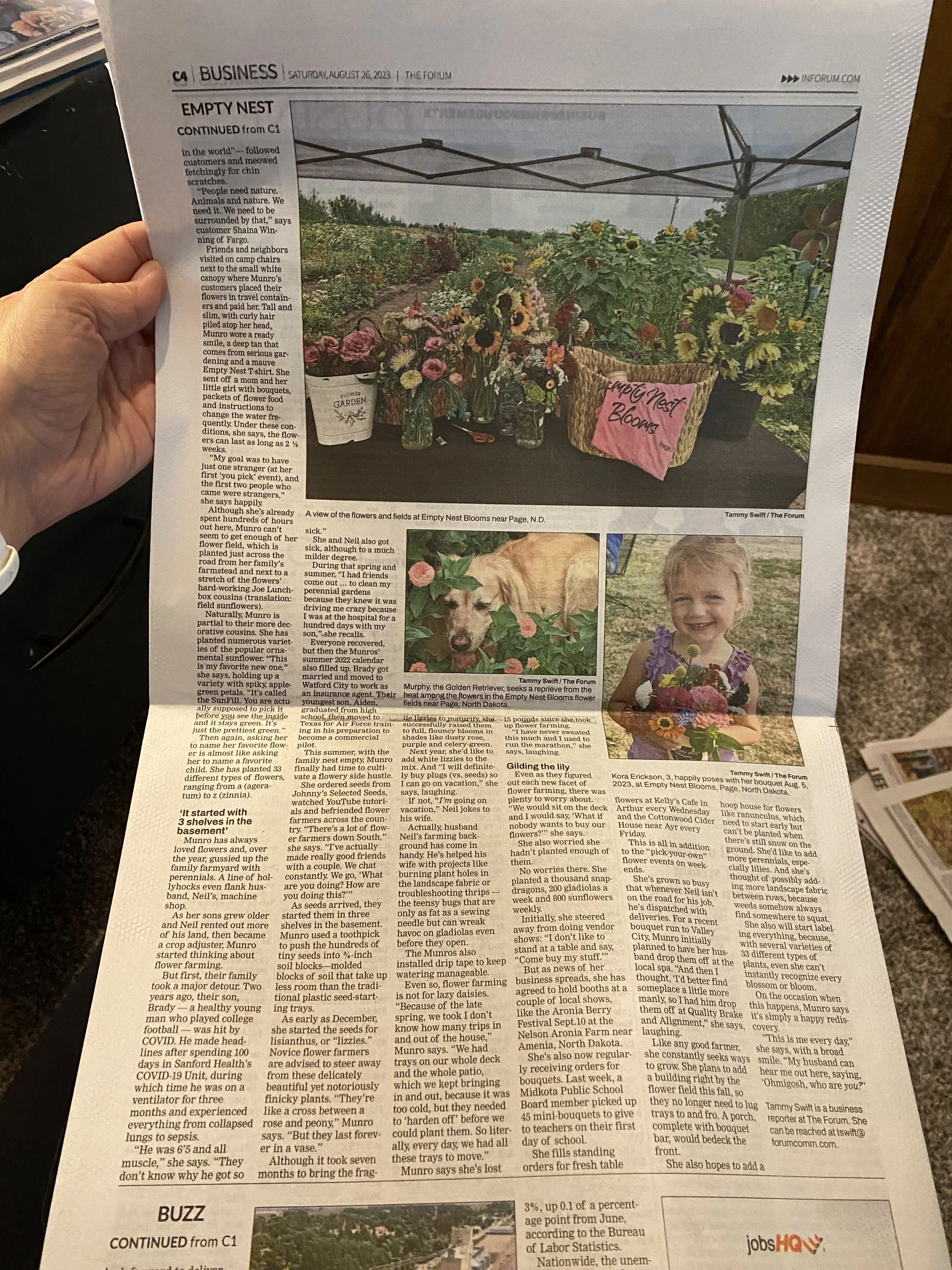 A newspaper page with a large photo of a flower display under a canopy, featuring sunflowers and other colorful blooms. The display includes a basket labeled "Empty Nest Blooms" and a small sign that reads "Garden."