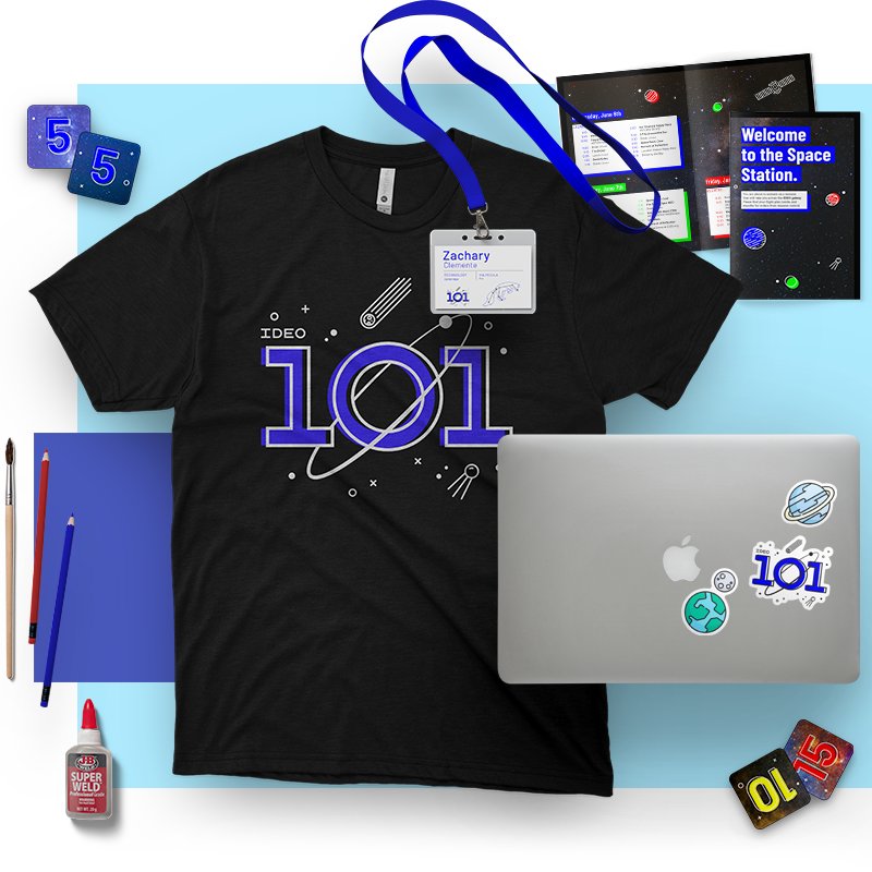 A space-themed workspace with a black t-shirt featuring '101' with space graphics, a name tag reading 'Zachary Clemente,' a closed laptop with space stickers, a blue notebook, pens, dice, a glue bottle, and space-themed brochures or posters.