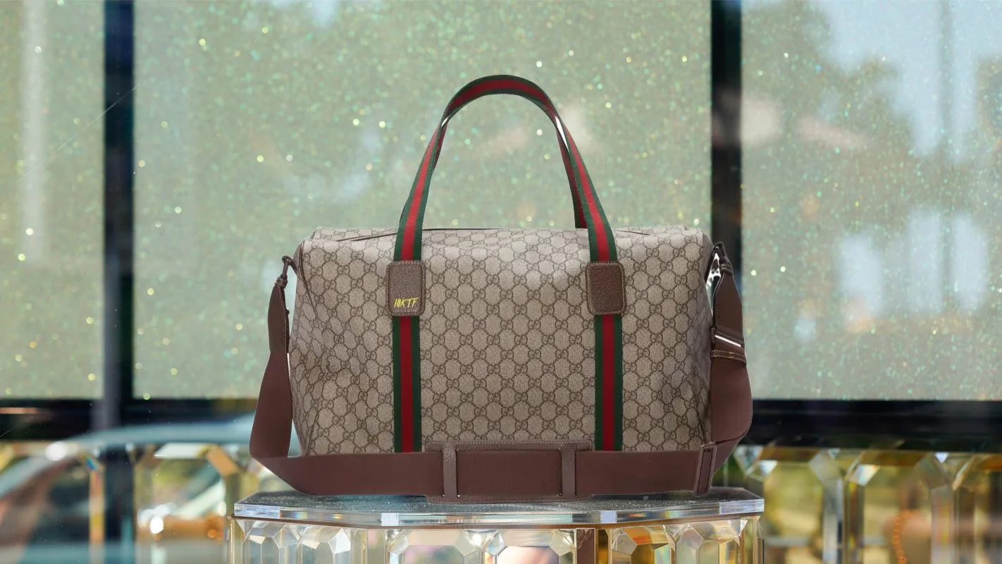 A beige Gucci duffel bag with green and red straps, displayed on a glass stand.