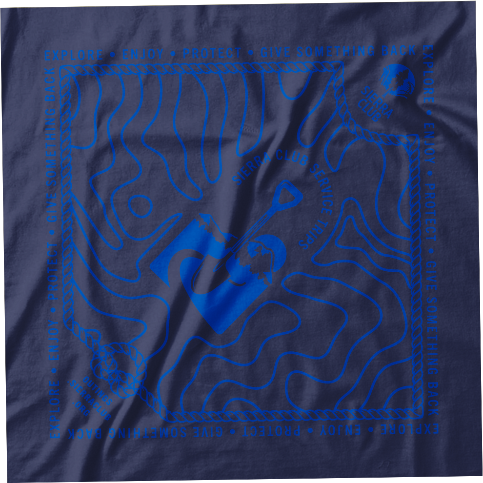 A dark blue cloth with a topographical map design in light blue, featuring a mountain range and a shovel. The border contains words like 'Explore,' 'Enjoy,' 'Protect,' 'Give Something Back,' repeated around the edges. The Sierra Club logo and commitments to service trips are also visible on the cloth.