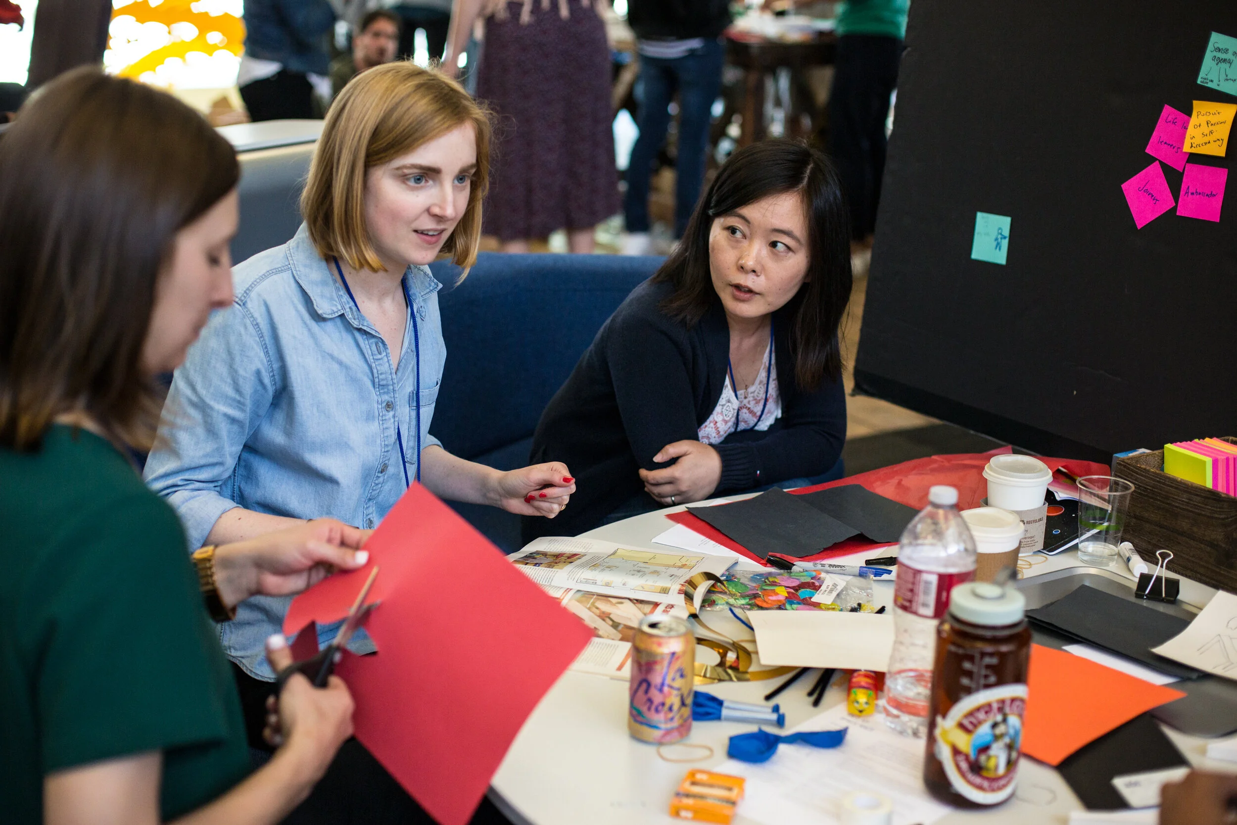 Three women sit at a cluttered table having a discussion. One woman is cutting red paper, another is speaking and gesturing, while the third woman in a dark sweater listens attentively. The table has various craft supplies, snacks, and drinks, with sticky notes on a blackboard behind them.