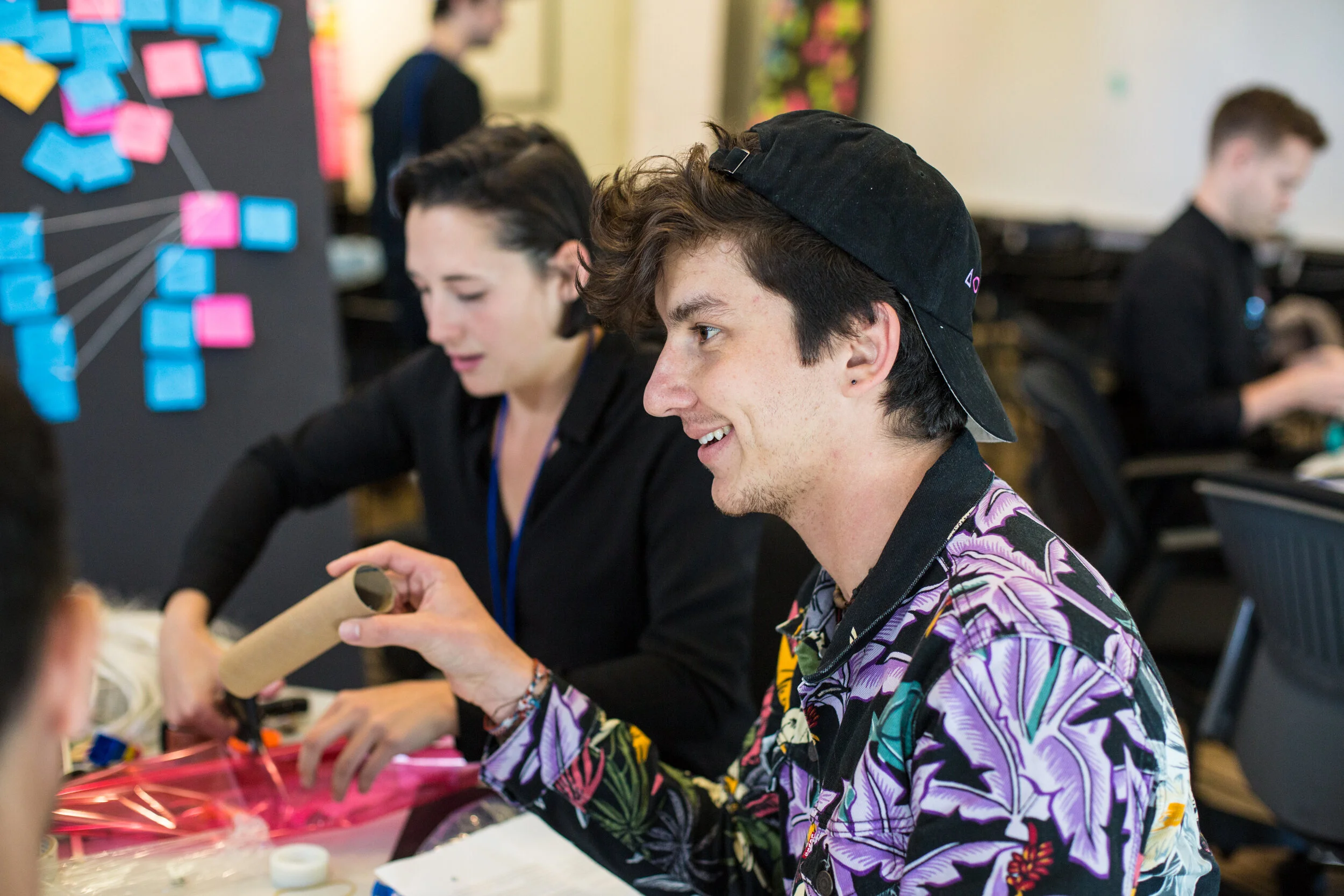 A young man wearing a black baseball cap and a colorful floral shirt smiling while working on a project with other people at a table in a classroom or workshop setting.