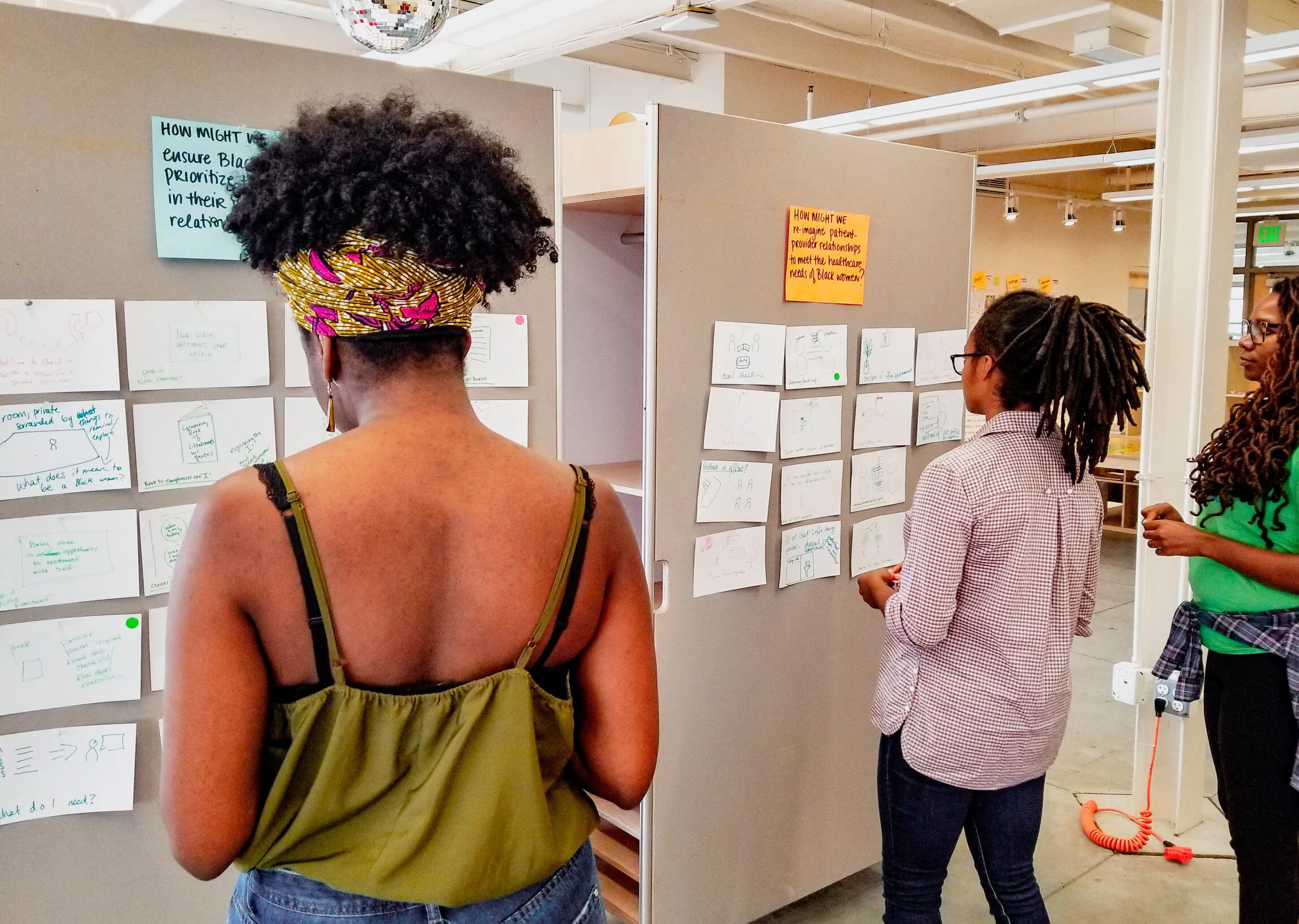 Three women standing in front of large grey display boards with notes, discussing ideas or projects in a well-lit indoor space.