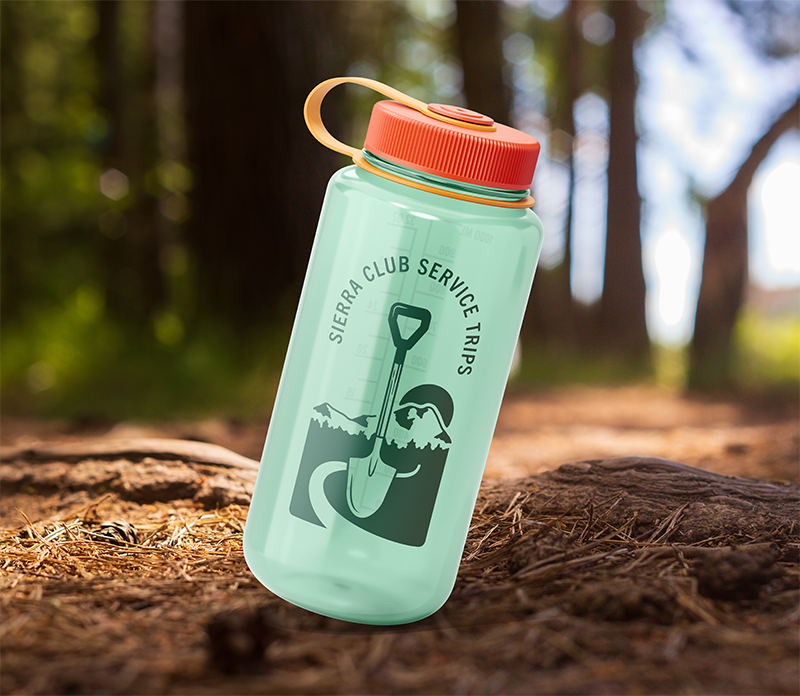 Green water bottle with orange lid and handle, featuring a graphic of a person with a shovel and the text 'Sierra Club Service Trips', placed on a forest trail.