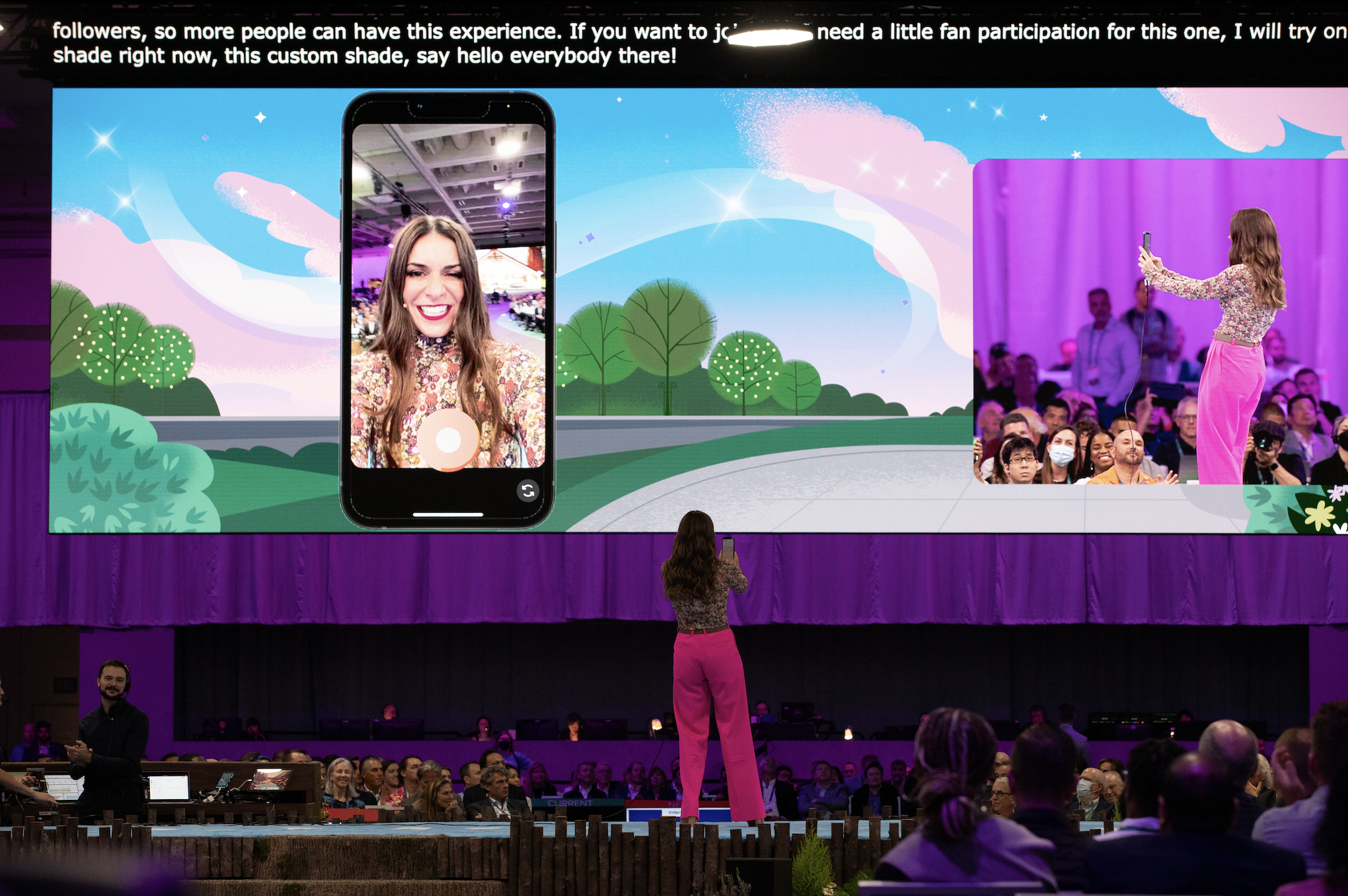 A woman stands on a stage in front of a large screen, taking a photo of the screen with her phone. The screen shows a presentation slide with a person smiling on a smartphone screen, overlayed on a colorful illustrated background of clouds and trees. The audience is seated in front of the stage, watching the presenter.