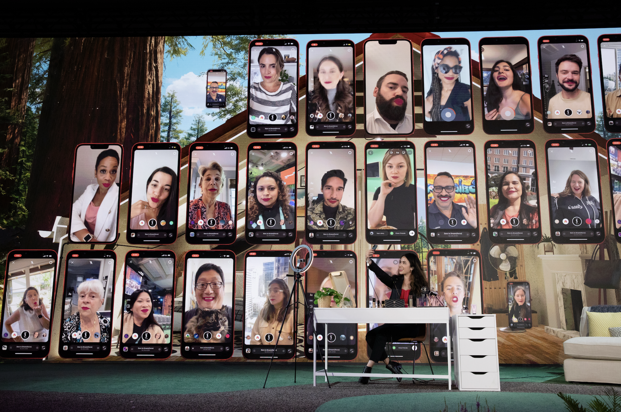 A woman sitting at a desk with a ring light and computer, surrounded by large screens displaying various people's face emojis, in a setting resembling a virtual meeting or video call collage