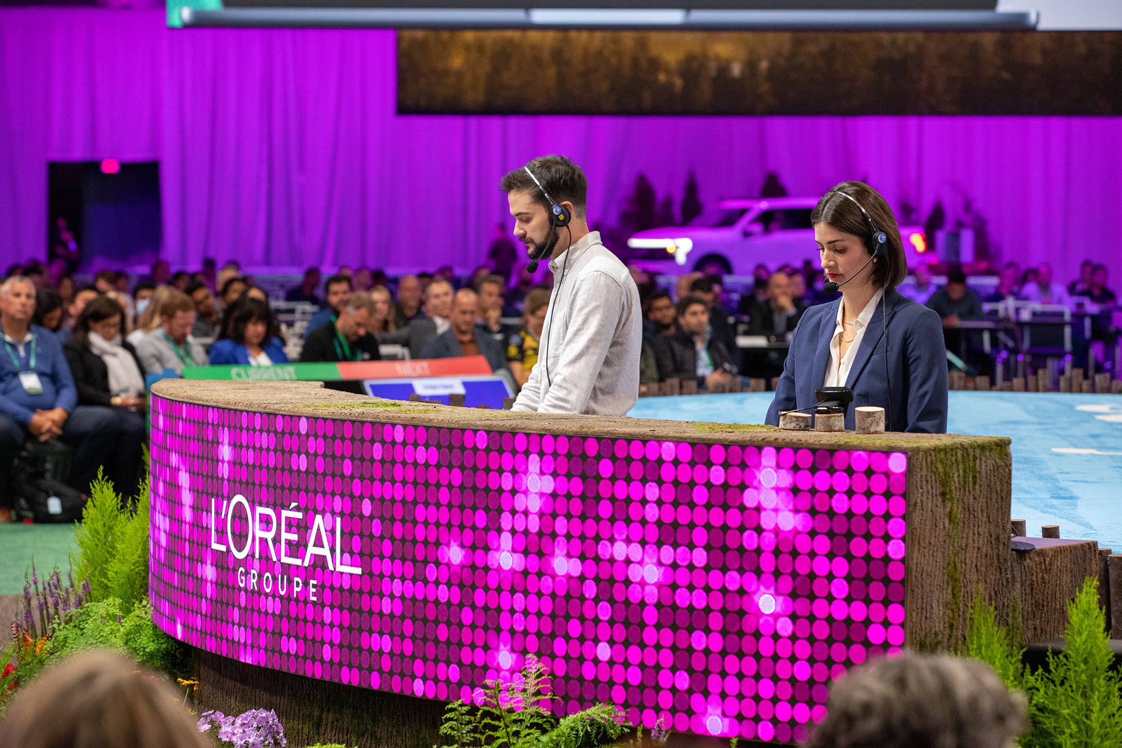 Two presenters at a conference, standing behind a curved digital display that reads "L'Oréal Groupe" with colorful LED lights, in front of a large audience seated in a hall with purple curtains and a car in the background.