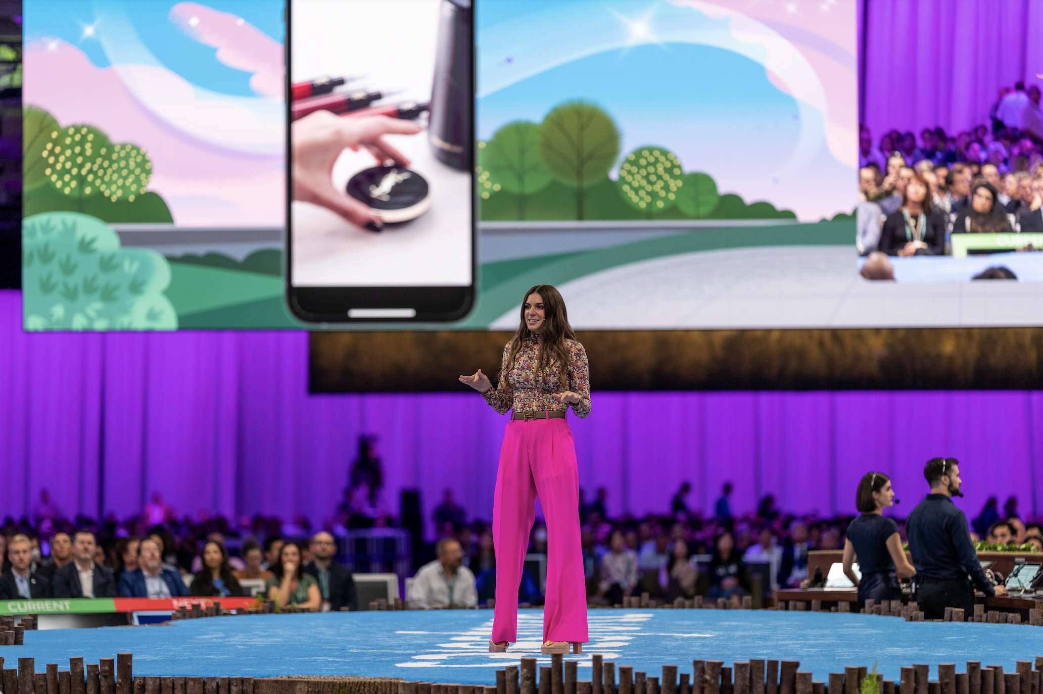 A woman in bright pink pants and a floral blouse is speaking on stage at a conference, with a large screen behind her displaying images of a hand holding a mobile device and a digital landscape. The audience is seated in front of her, with purple curtains and stage lighting.