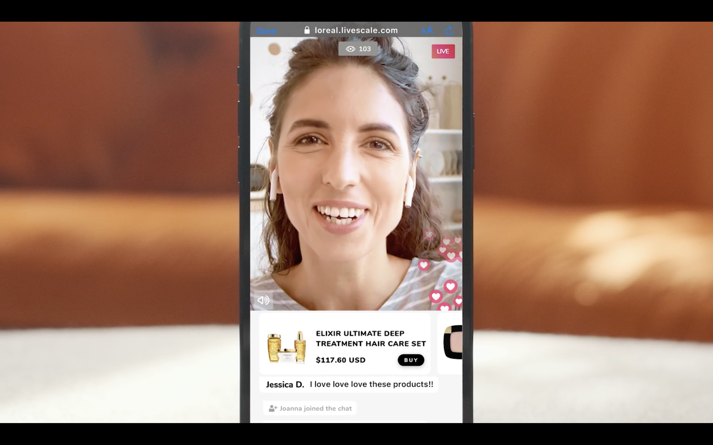 A woman on a live video shopping stream smiling and wearing wireless earbuds, with product information for a hair care set displayed at the bottom.