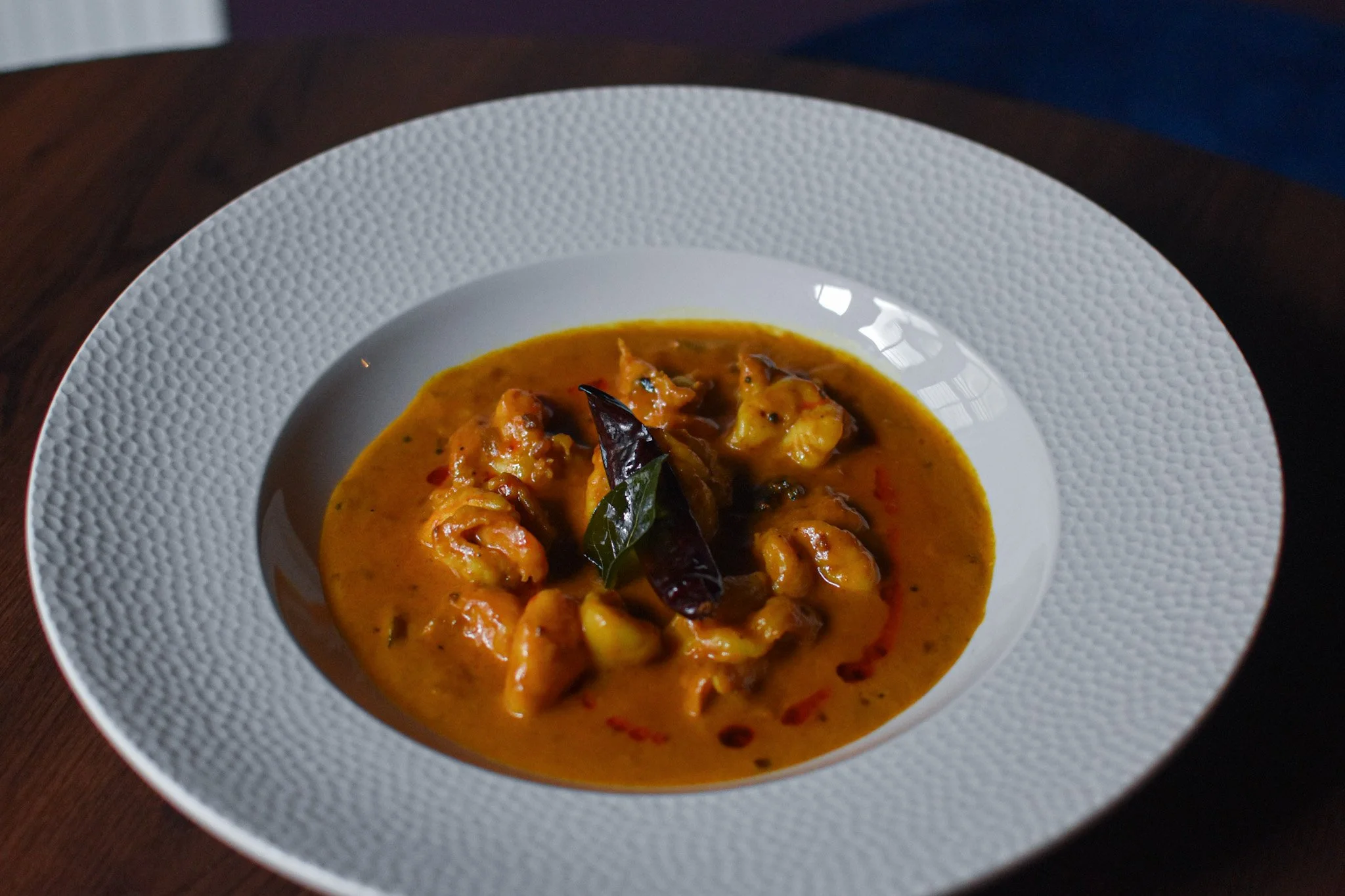 A bowl of yellow curry with chunks of meat or vegetables, garnished with dark leaves, served in a white textured bowl on a dark wooden surface.