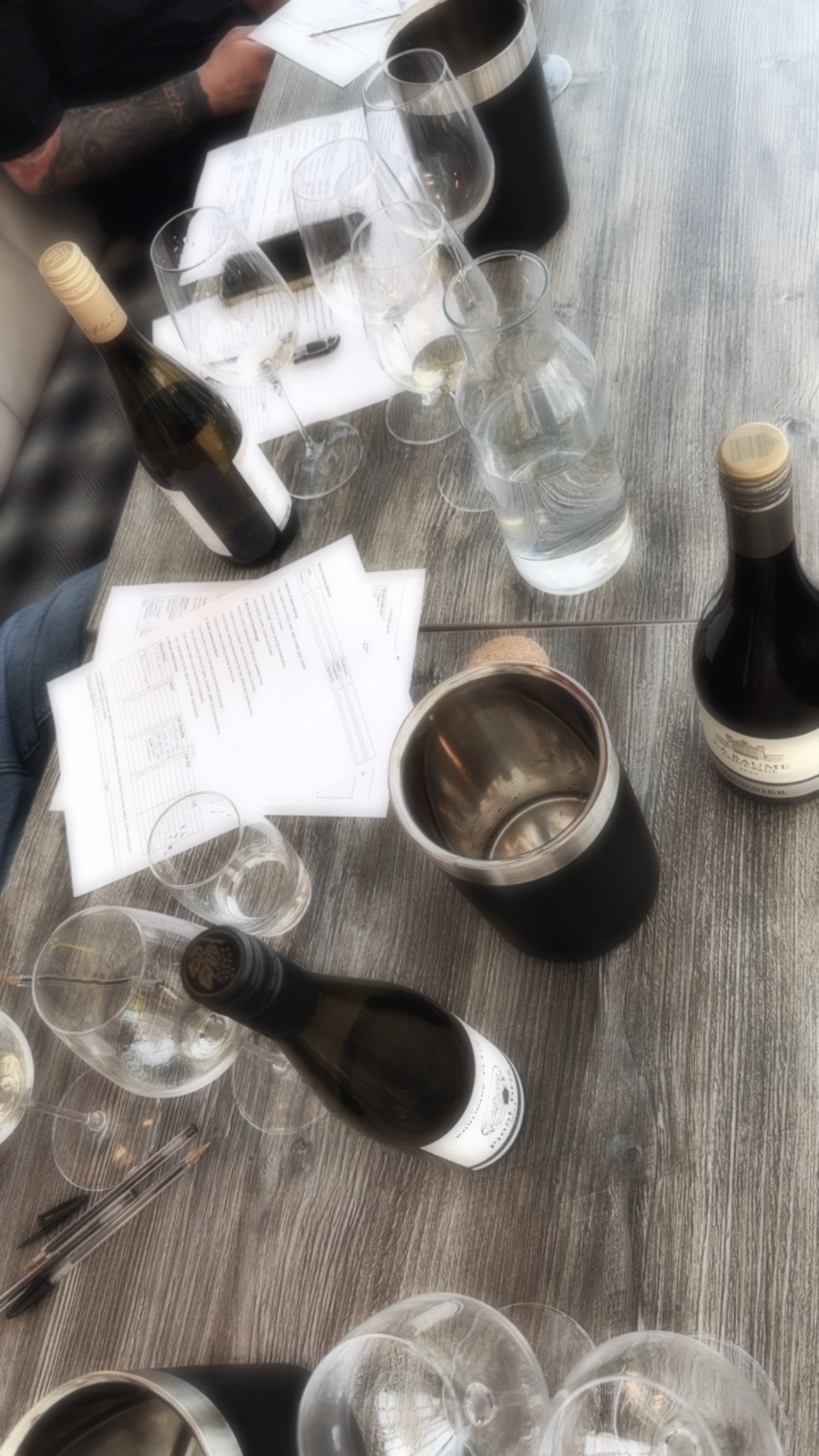 Table with multiple wine bottles, empty wine glasses, a water pitcher, and printed sheets of paper. A person with a tattooed arm is visible sitting at the table.