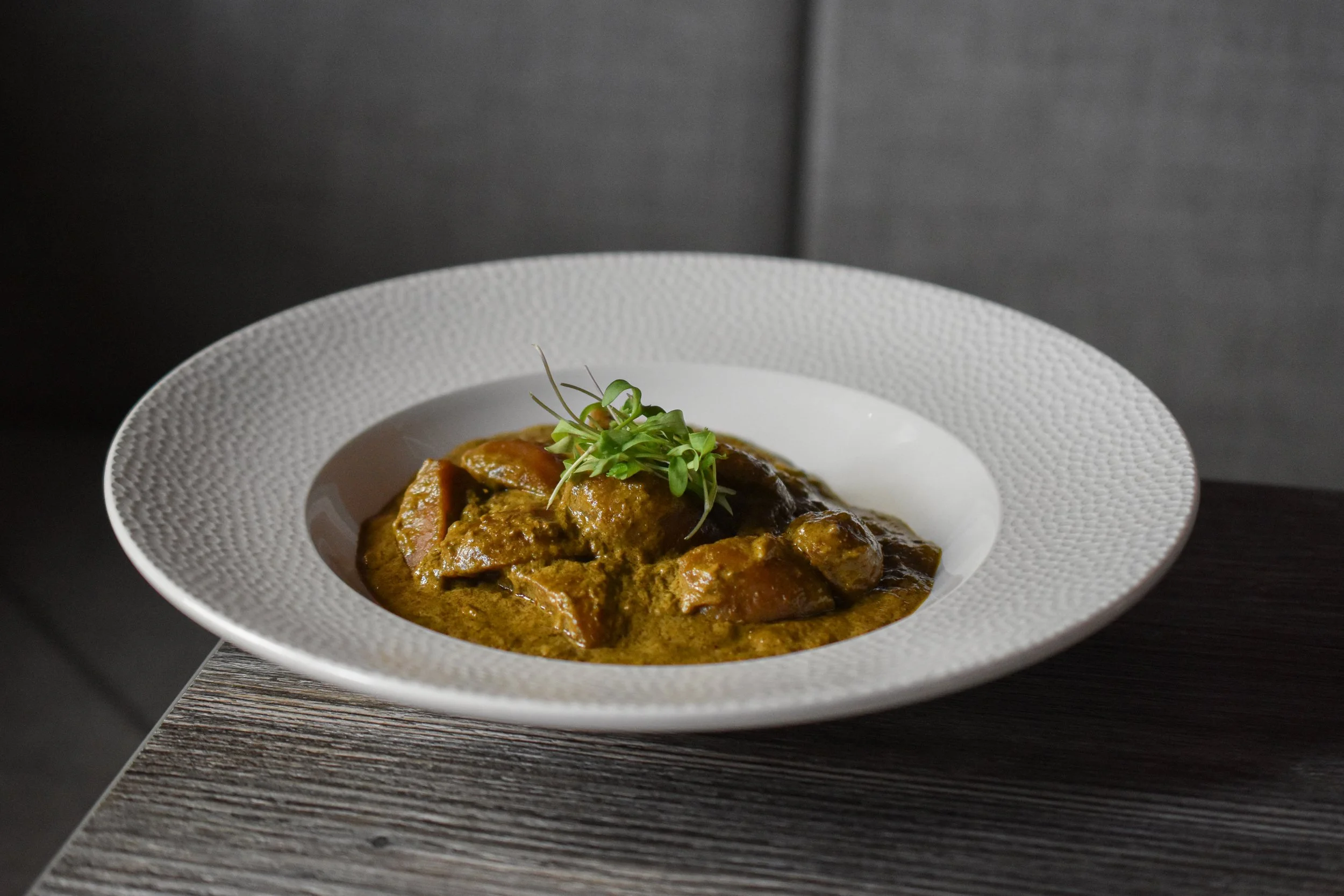 A white textured bowl containing a rich, brownish-golden food dish, garnished with sprouts, placed on a dark wooden table against a gray background.