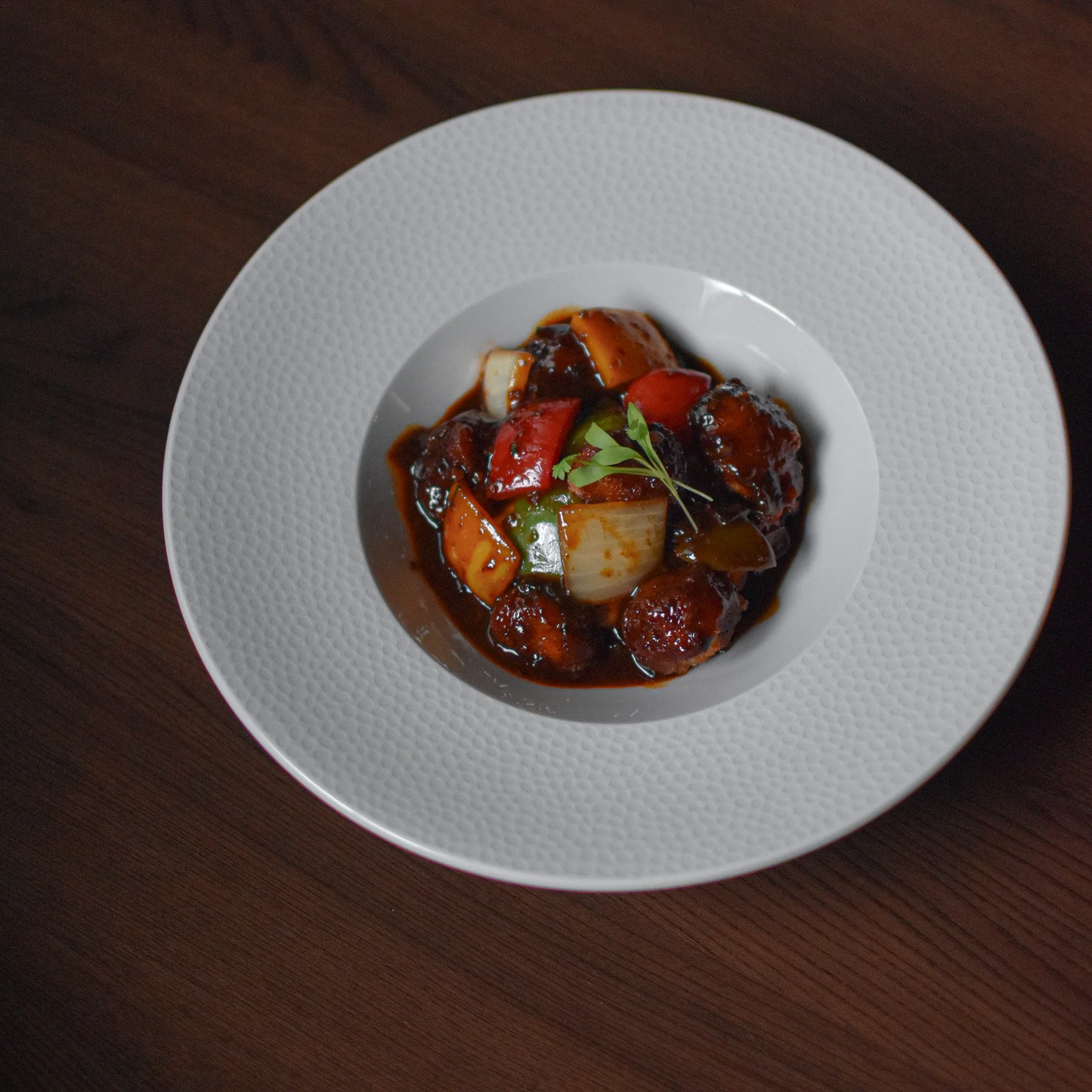 A white textured plate with a serving of sweet and sour pork with red and green bell peppers, pineapple chunks, and garnished with a small sprig of parsley, on a dark wooden table.