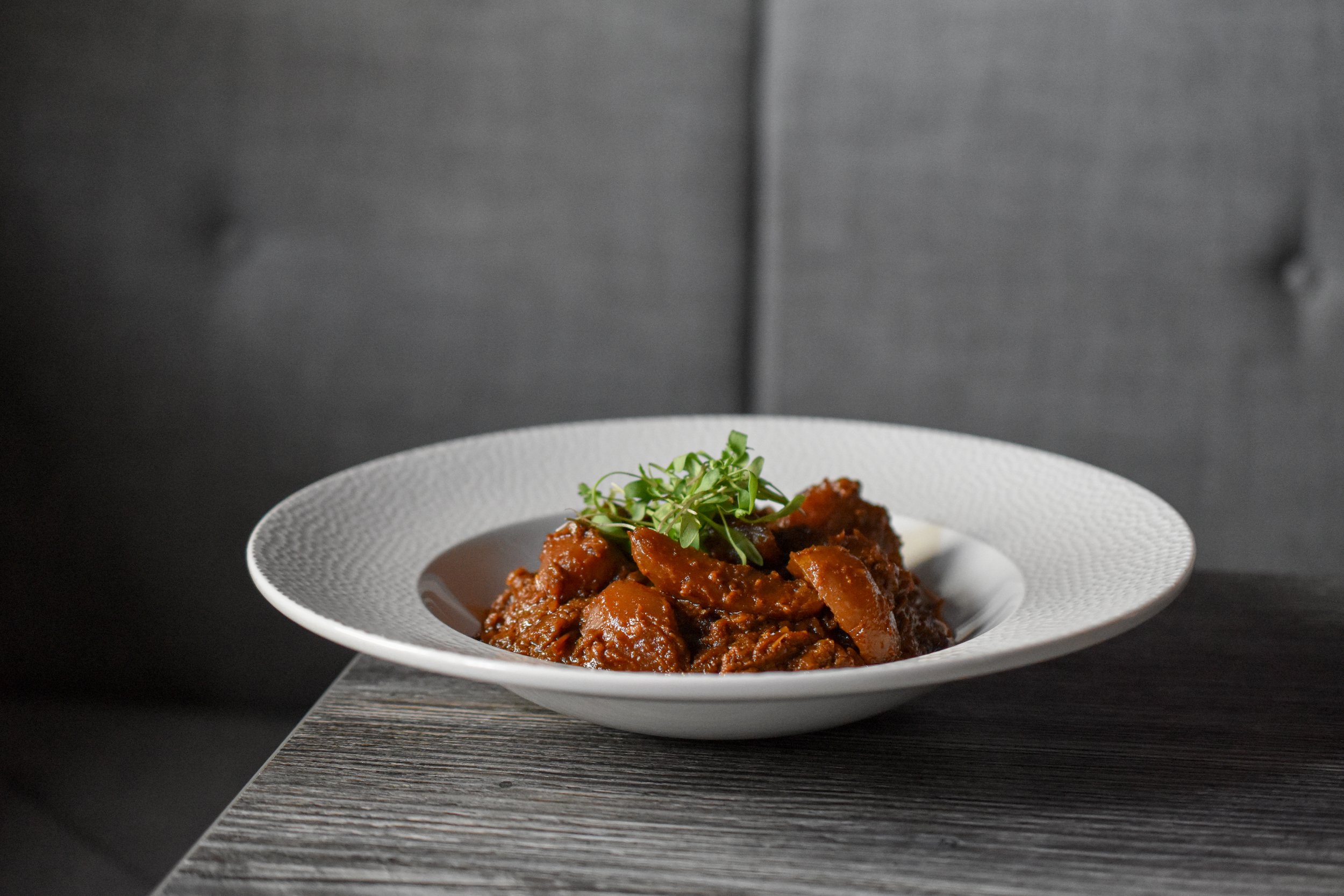 A white textured bowl filled with a meat stew garnished with green microgreens, set against a dark background.