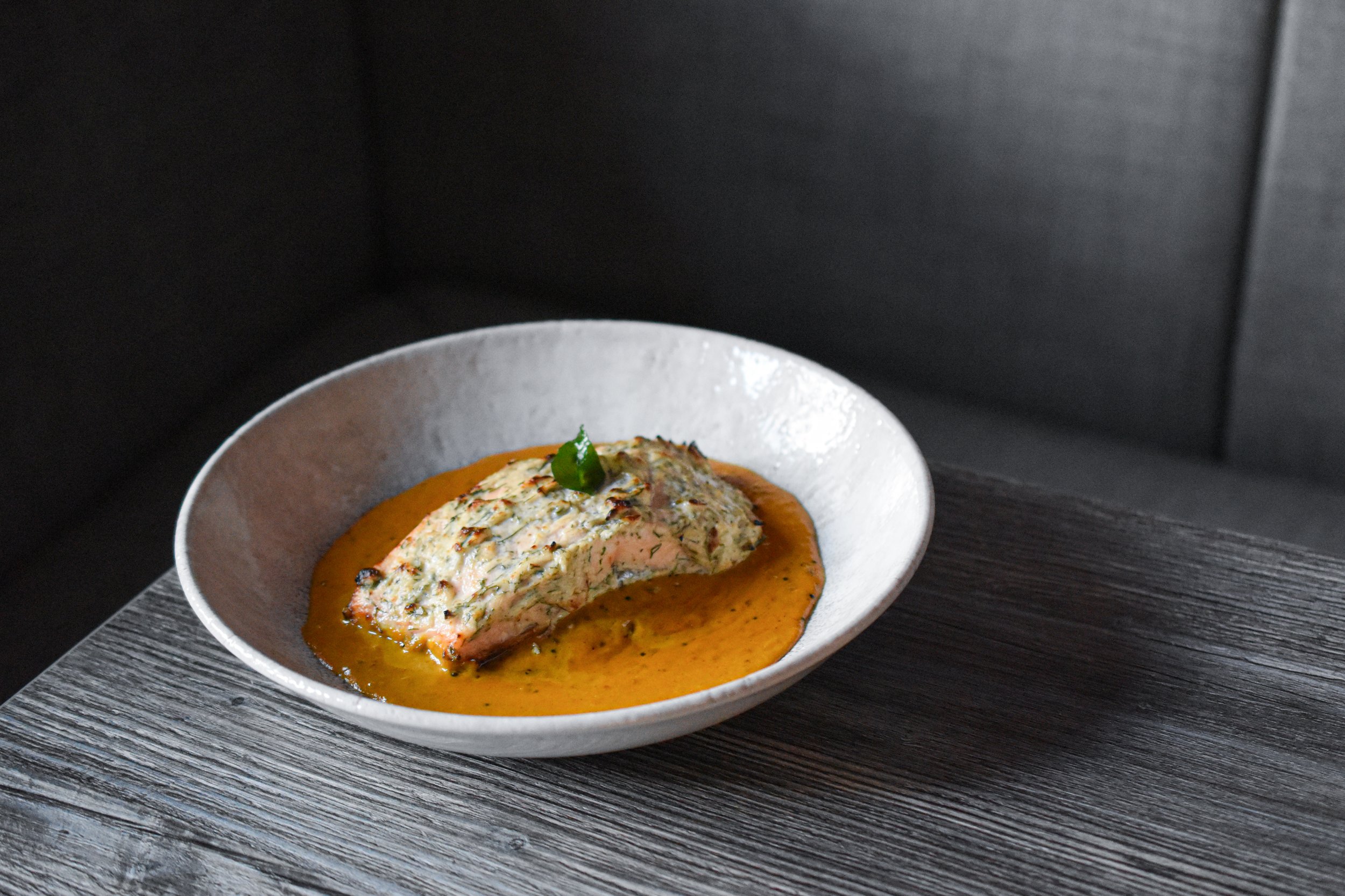 Salmon Mappas at Nomi Porthmadog, a modern Indian salmon dish with creamy herb crust, coconut based sauce and fresh garnish, served on a black wooden table.