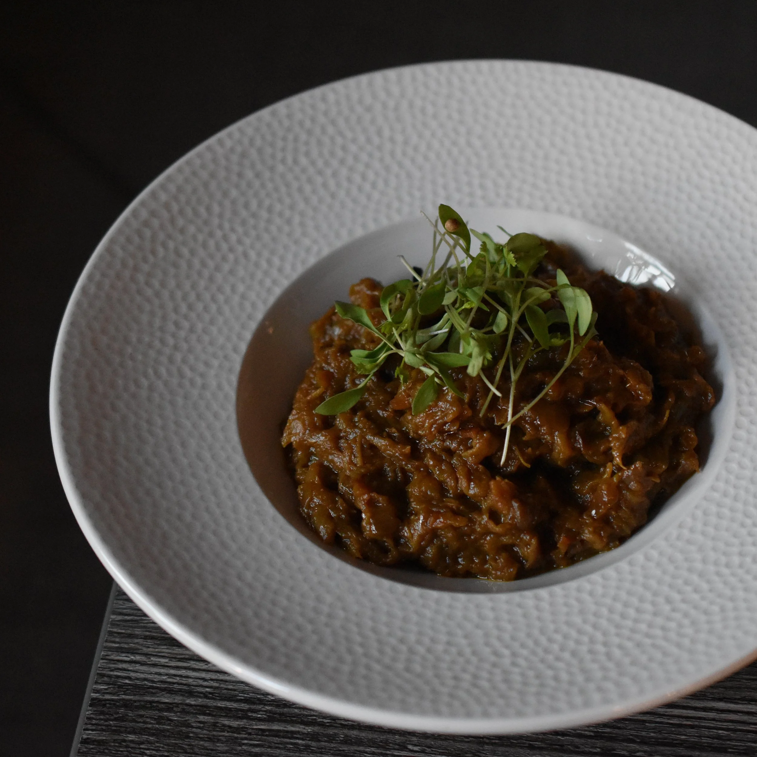 Close-up of a textured white plate with a portion of dark brown food topped with microgreens, placed on a dark wooden surface.