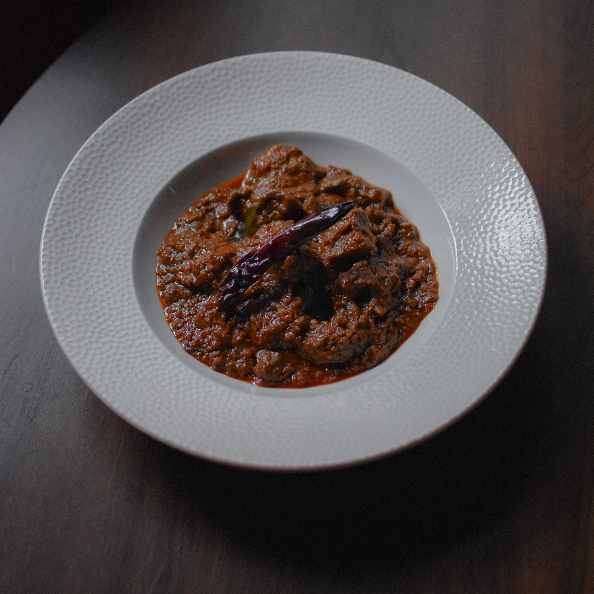 Close-up of cooked meat in a white textured plate, garnished with dried red chili peppers, on a black surface.