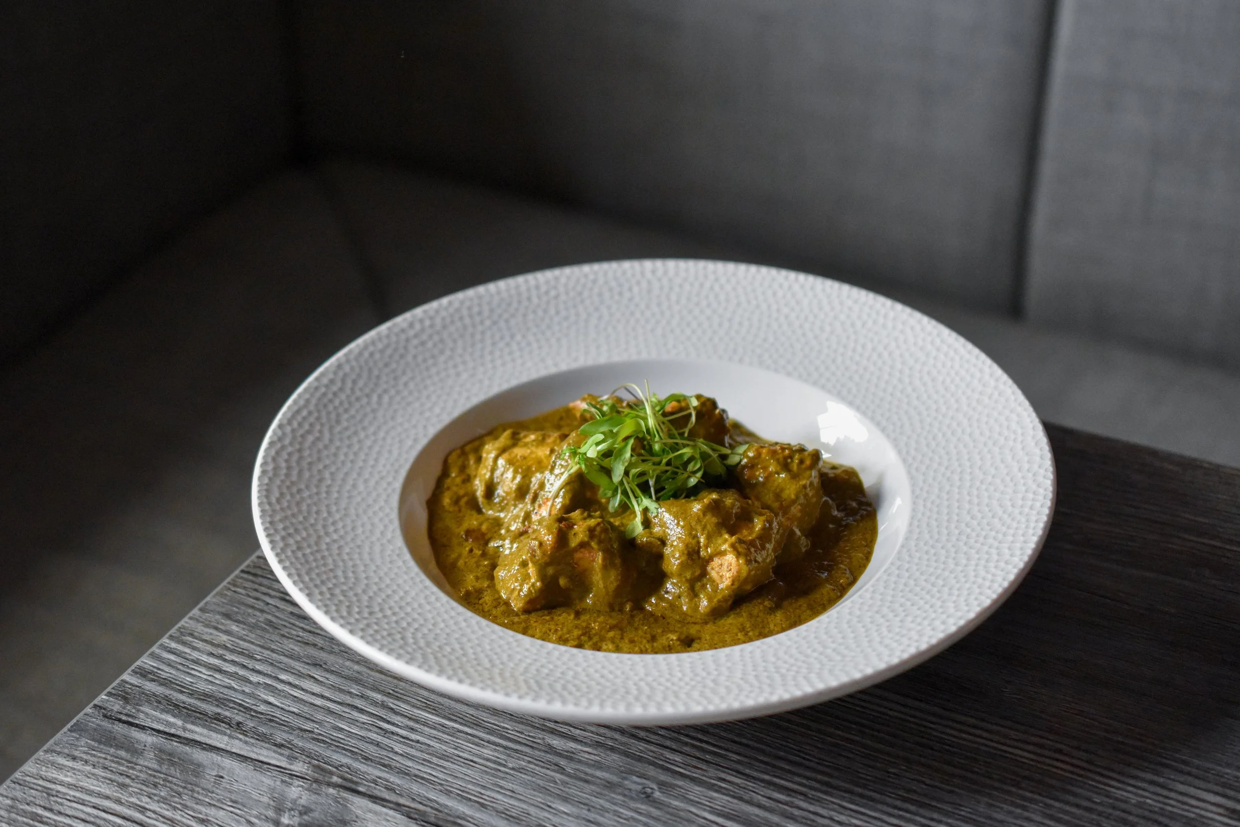 A white textured bowl filled with a curry dish topped with microgreens, placed on a wooden table with a dark background.