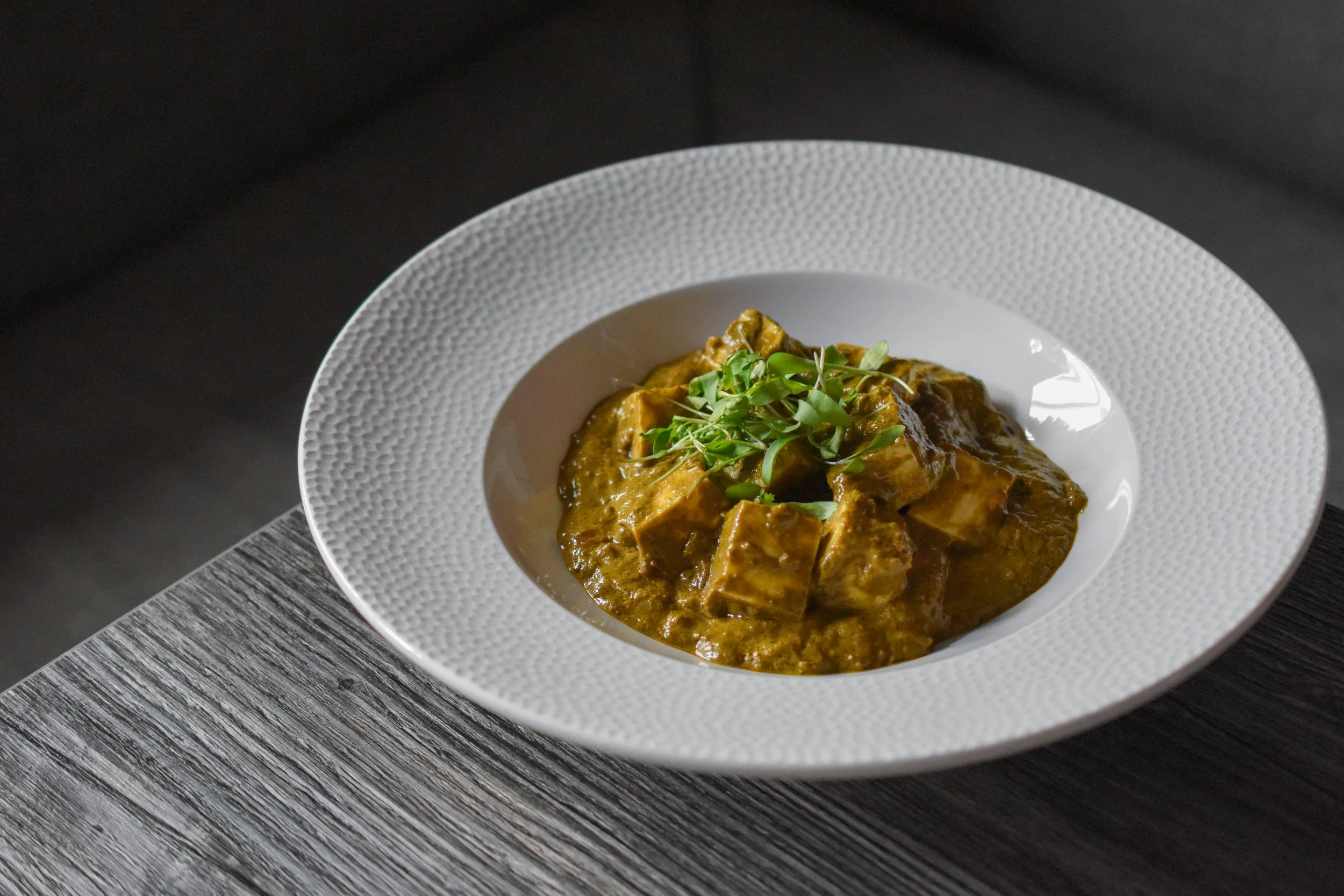 A white bowl with a textured rim contains a serving of curry topped with fresh greens, placed on a dark wooden surface.