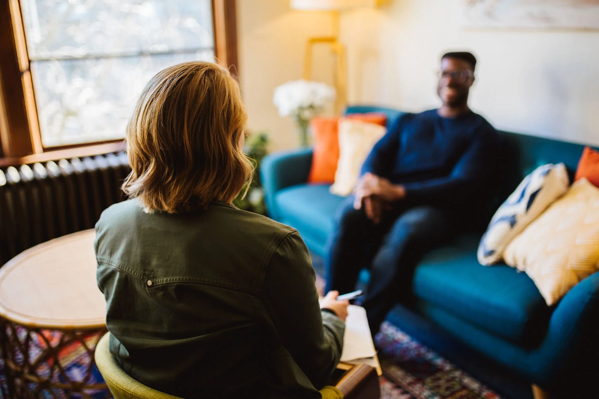 Therapist seated in therapy office, holding a notepad and pen, facing a midlife man sitting on a blue sofa with colorful pillows. The sacred space in Bellingham, WA has a window with daylight streaming through.