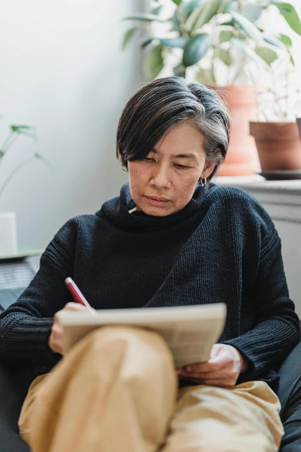 A woman in her 50s with short dark hair and earrings, wearing a black sweater, sitting on a couch with plants in the background, writing in her dream journal.