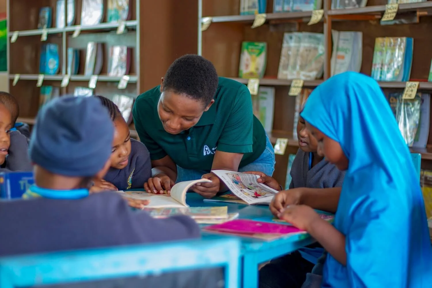 An Amani staff member leans over a table to read with a small group of children in a well-stocked library, engaging them in a hands-on literacy session.