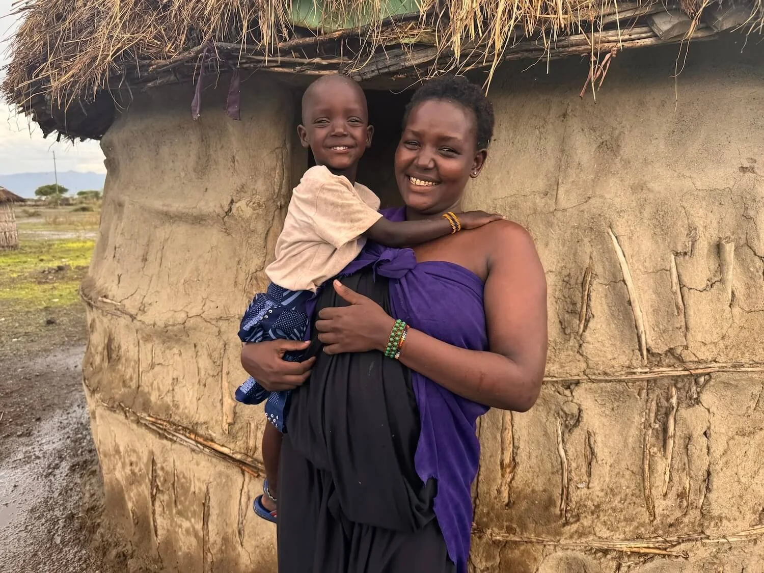 A young mother smiles warmly while holding her child outside a thatched mud home in a rural Tanzanian village, with an open landscape visible in the background.
