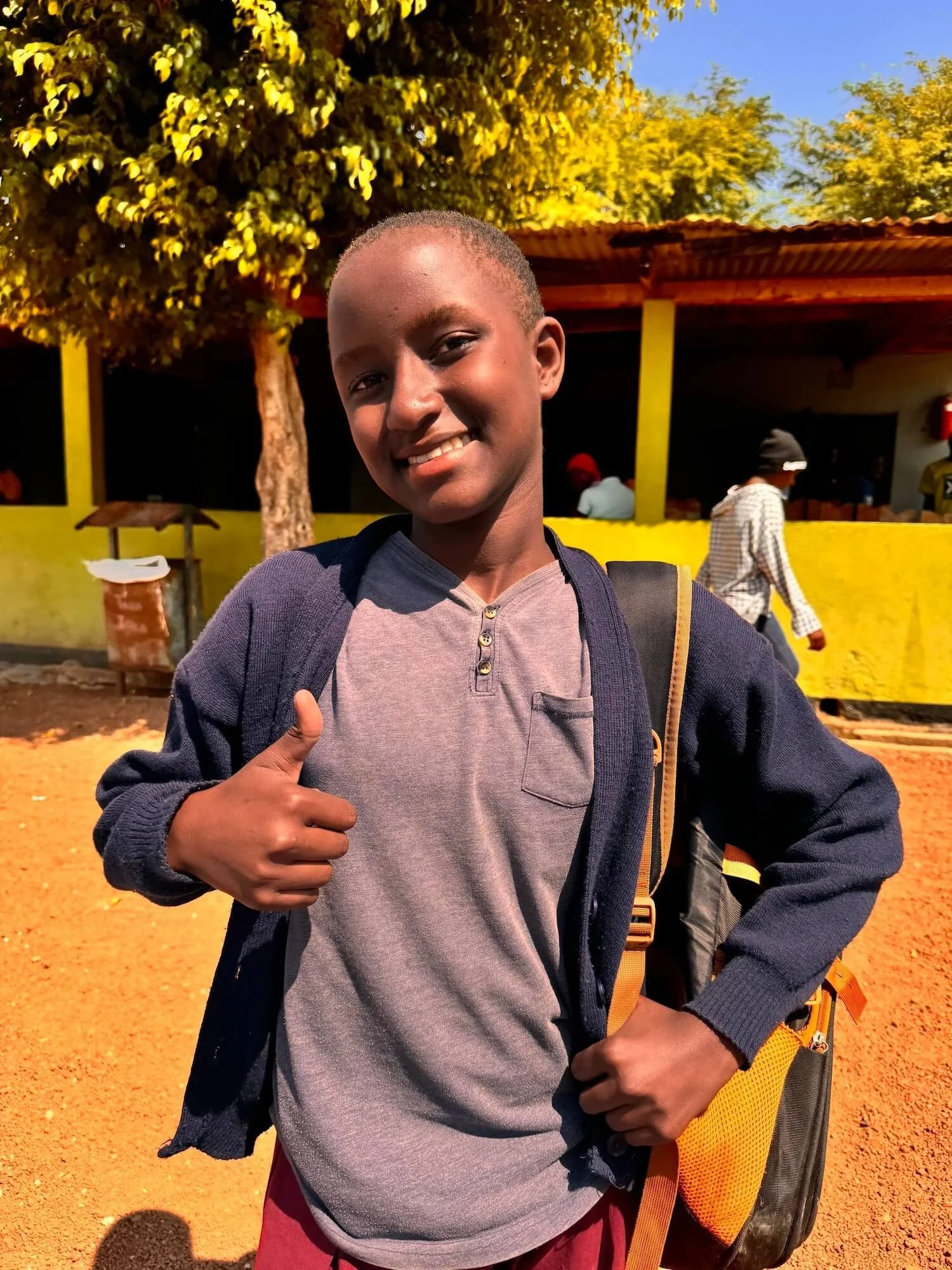 A young boy gives a thumbs up and smiles confidently in a sunny school courtyard in Tanzania, wearing a backpack and school uniform sweater.