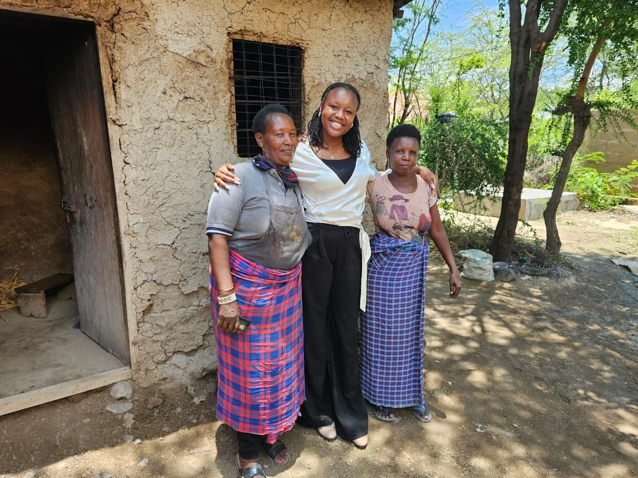A staff member from Amani smiles and poses with two community women wearing traditional plaid wraps outside a mud-brick home in rural Tanzania.