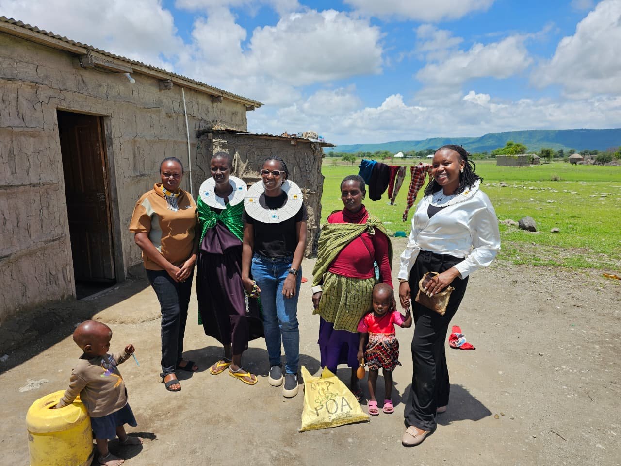 A group of Amani staff members stand together with a local mother and her young children outside a rural home in Tanzania, with rolling green hills in the background.