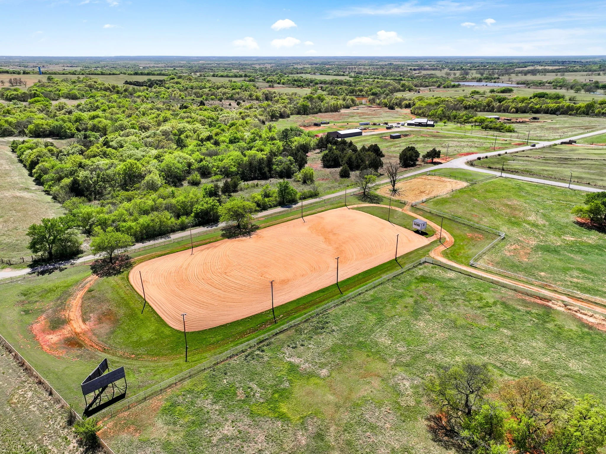 An aerial view of a baseball field under construction with a dirt infield, surrounded by a grassy outfield and trees, with some houses and roads in the background.