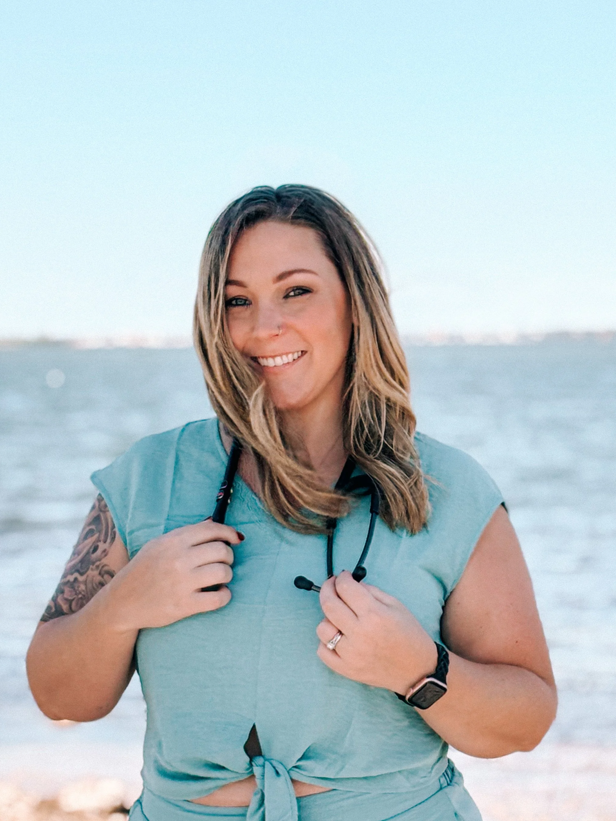 A smiling woman with shoulder-length brown hair holding a pair of black earphones, standing on a beach with the ocean and sky in the background.
