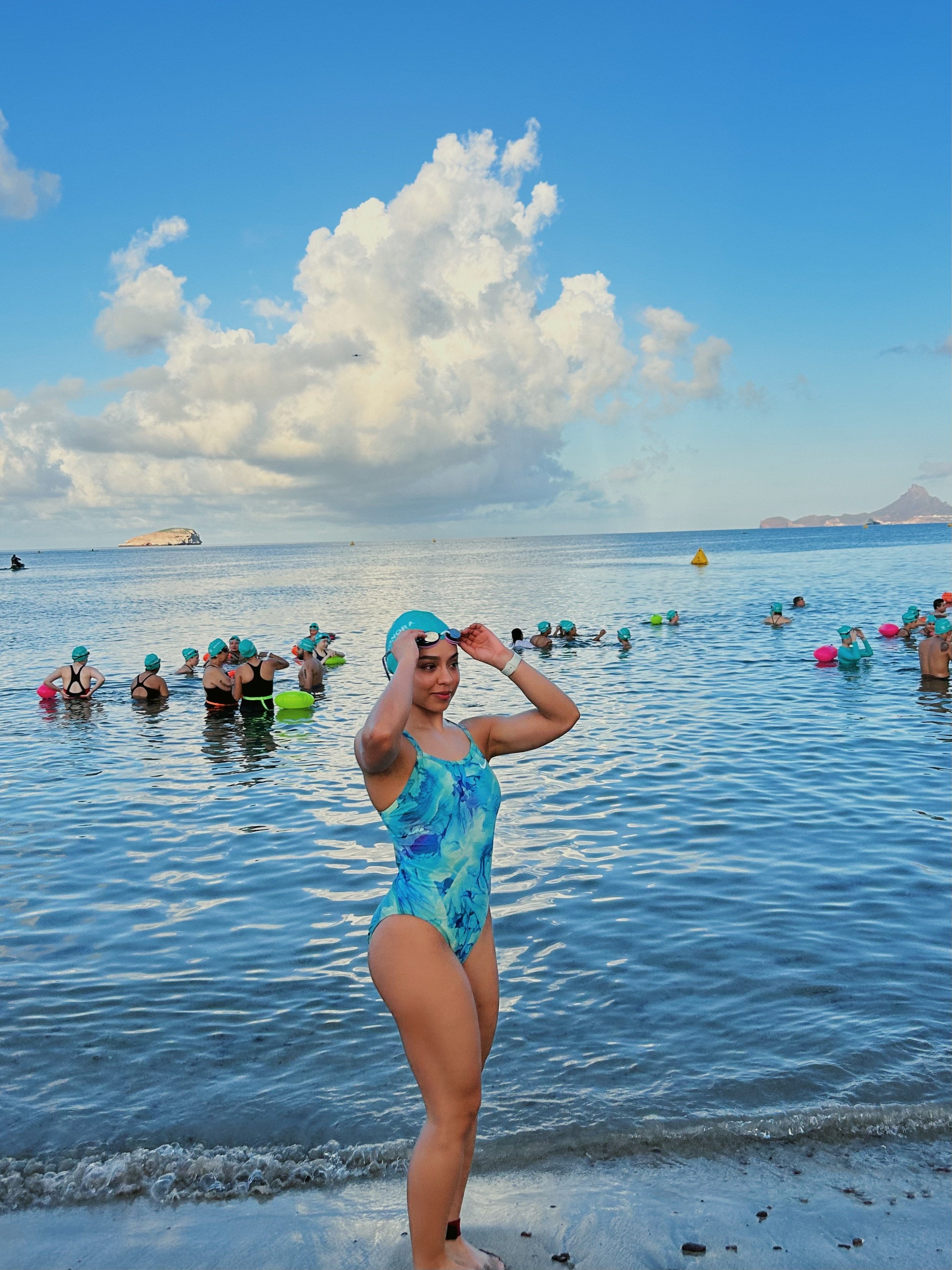 Una mujer con gorra y traje de baño de escote en V, de pie en la orilla del mar, ajustándose la gorra, con muchas personas en el agua y huestes en el fondo bajo un cielo nublado.