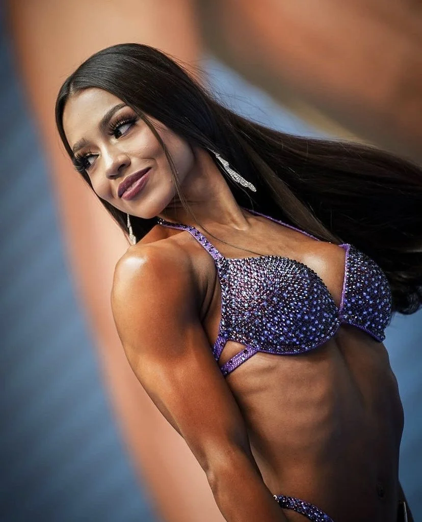 Mujer en vestido de competencia de culturismo, mostrando músculos tonificados, con piel bronceada y maquillaje, usando joyería y cabello largo y liso.