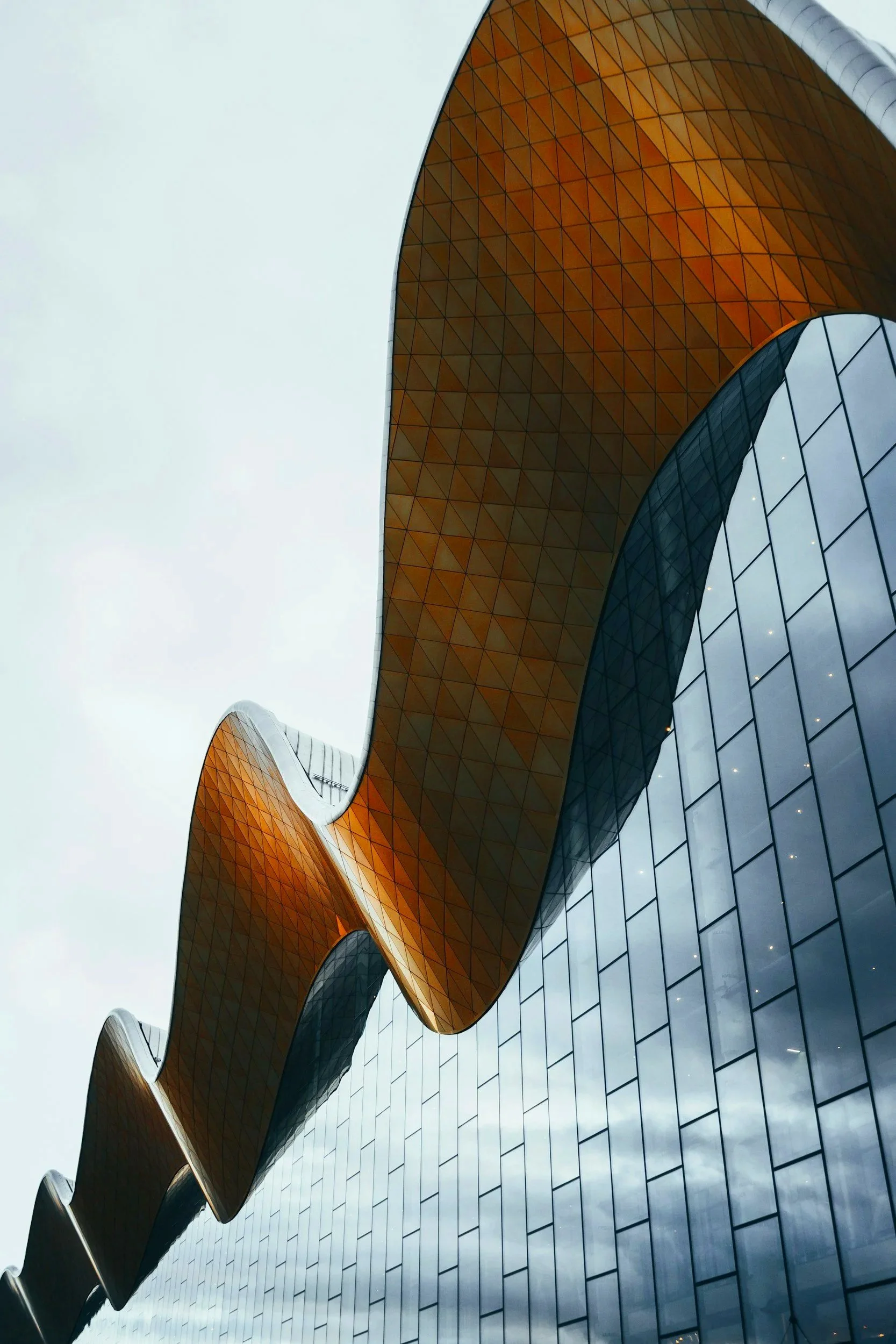 Modern building with undulating orange and black metal roof and glass facade, seen from below against a cloudy sky.