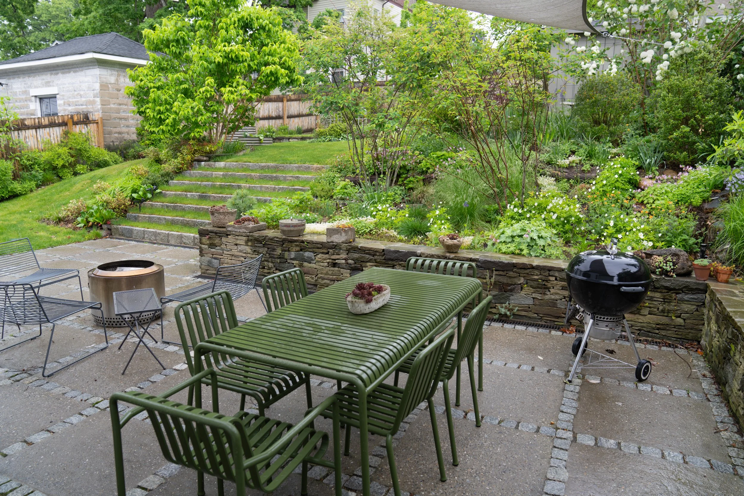 A backyard patio with green metal furniture, a charcoal grill, a stone wall, and a lush garden with trees, plants, and flowers. Stone steps lead up to a grassy area with more trees and bushes.
